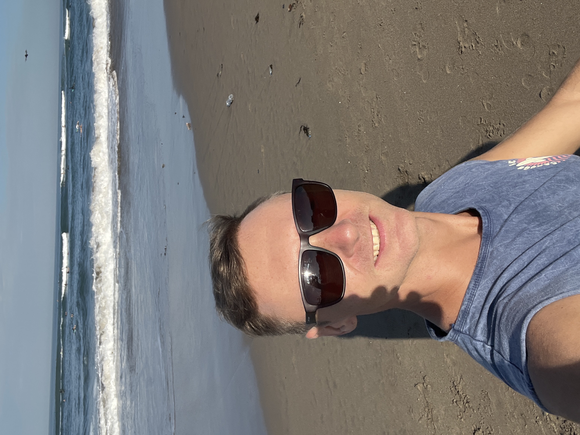 A man wearing sunglasses and a blue t-shirt is taking a selfie on a beach. The ocean is visible in the background with waves and several surfers. The beach is sandy and appears to have some litter on it, including plastic bottles. The sky is clear and the sun is shining. The overall scene depicts a casual day at the beach in North Kuta, Indonesia.