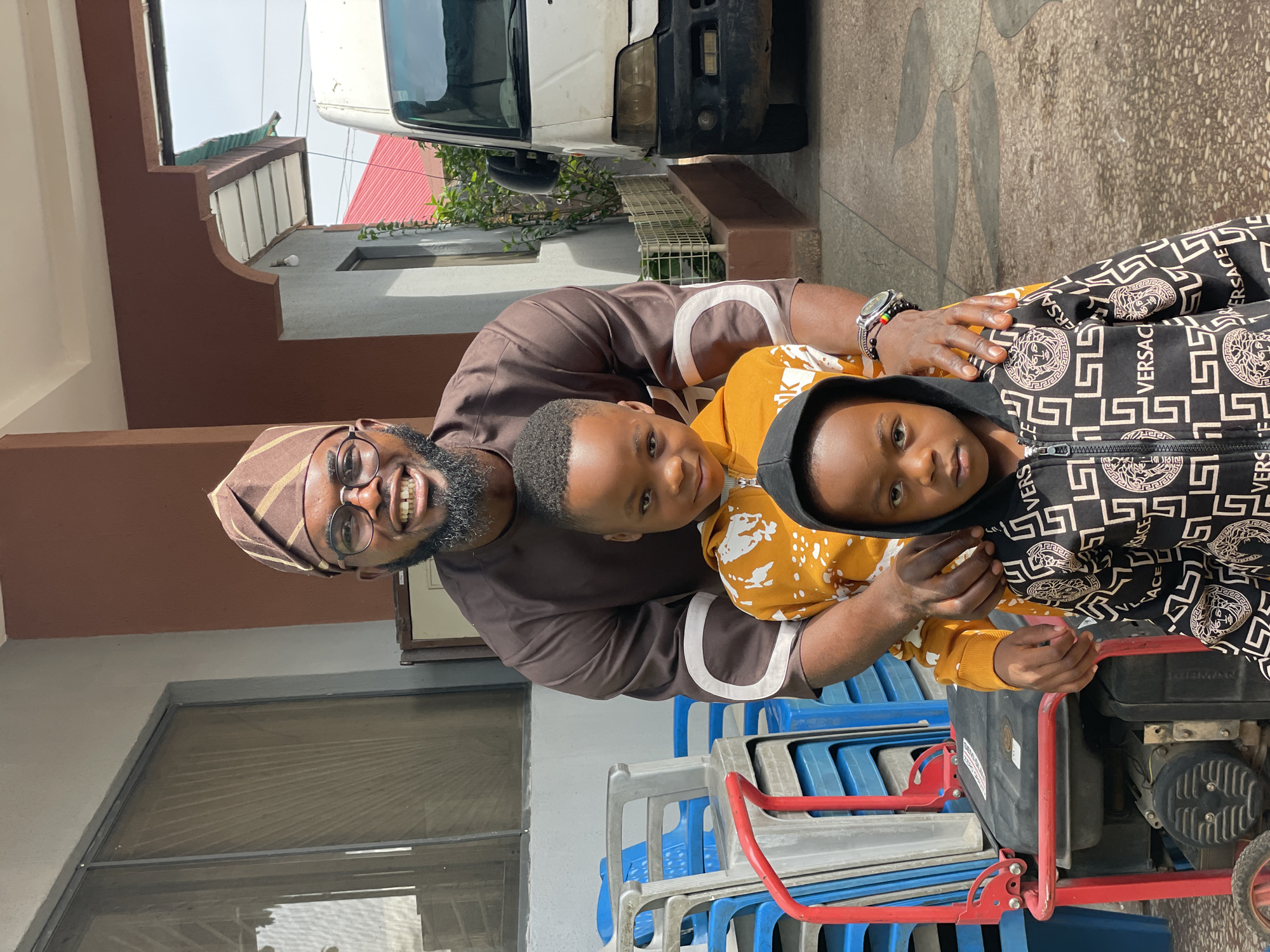 A man wearing a brown traditional outfit and cap, glasses, and a beard smiles at the camera. He has his arms around two young boys. The boy in the middle wears a yellow hoodie with a white abstract pattern, and the boy in front wears a black hoodie with a repeating gold pattern and the word "VERSACE" visible. They are standing outdoors in what appears to be a residential area in Jos, Nigeria. In the background, there are stacked blue and grey plastic chairs, a red and black generator, and a white van. The ground is paved with patterned concrete. The wall behind the man is painted a dark brown, and the building has a white-trimmed overhang. The sky is visible above.