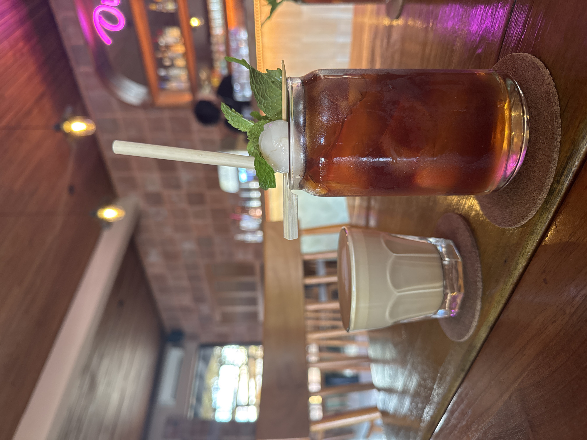The image shows two beverages served in glasses on a wooden table. The first beverage is a dark liquid, possibly iced tea or coffee, garnished with mint leaves and a lychee. A natural straw is placed in the glass. The second beverage is a lighter-colored, creamy liquid, potentially milk or a latte. The drinks are resting on round coasters. The background reveals a bar setting with bottles and a neon sign, suggesting the location is a cafe or restaurant. The location is the city of Denpasar, Indonesia.