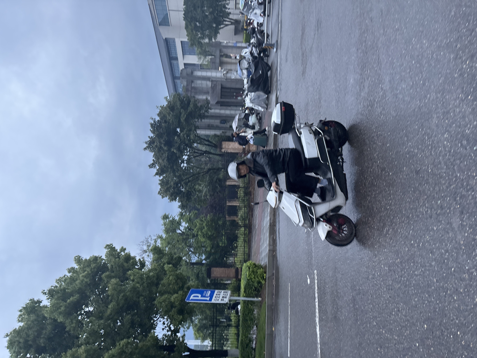 A medium shot captures a person riding a white motor scooter on a wet street in Jiangjin, China. The rider wears a helmet and dark clothing. The scooter is positioned on the right side of the frame, angled slightly away from the camera. The street appears to be damp from recent rain. In the background, trees line the street and a building is visible. Other parked scooters are seen lined up next to the building. A blue parking sign with Chinese characters is on the left side of the road, near the scooter. The sky is overcast.