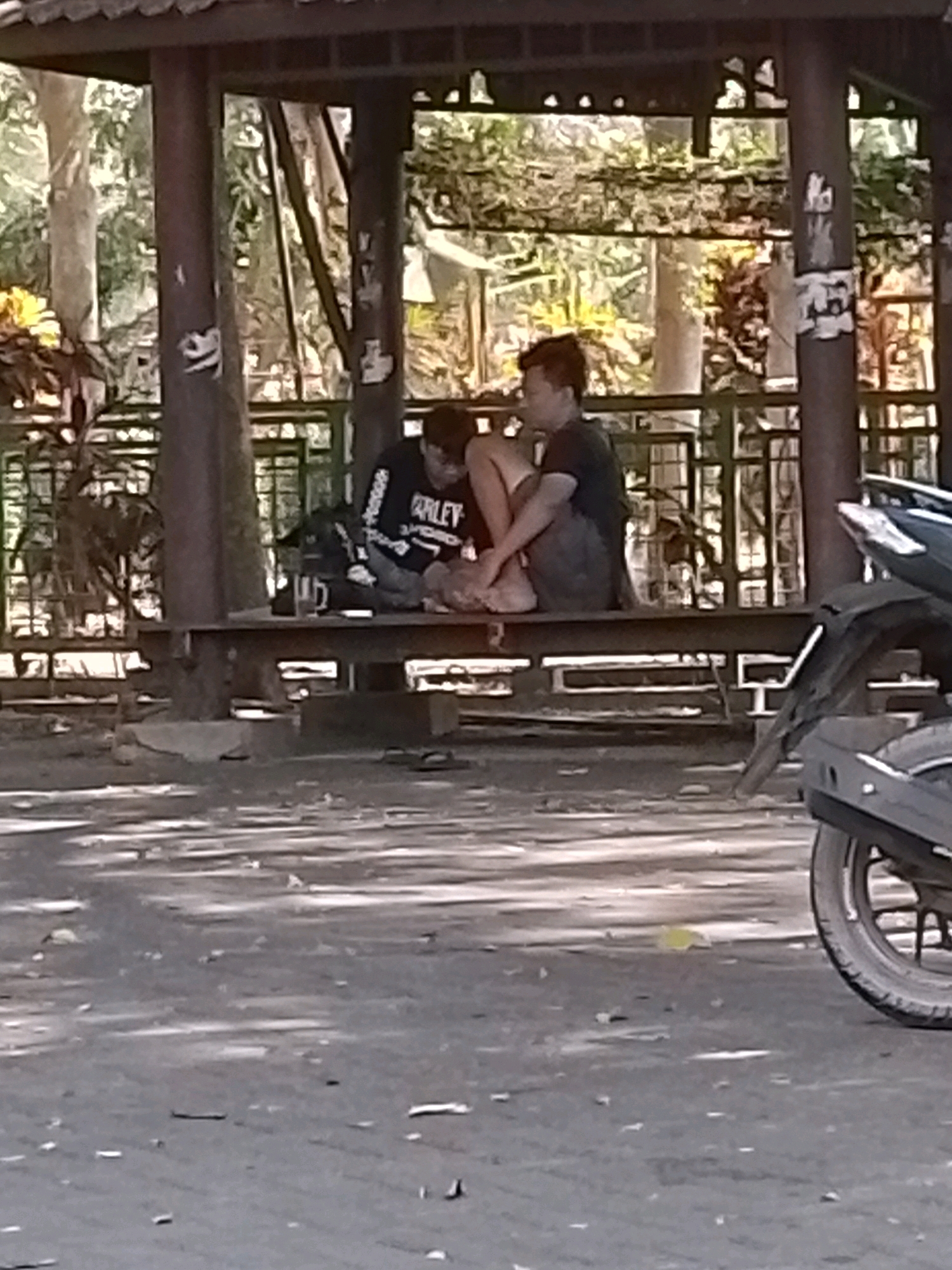 The image depicts a shaded outdoor seating area, likely a gazebo or pavilion, located in a park or public space in Wonosari, Indonesia. Two young men are seated on a wooden bench within the structure. The man on the left wears a dark long-sleeved shirt with white lettering that appears to say "HARLEY" and dark pants. He is seated with his legs crossed. The man on the right wears a dark t-shirt and shorts. He is sitting with his legs bent and crossed, leaning his head on his hand. A dark backpack is placed on the bench between the two men. The gazebo has dark brown pillars and a slatted roof. Behind the men, a metal fence with vertical bars is visible, with lush green foliage and trees beyond. The ground in front of the gazebo is paved with asphalt, and a portion of a dark-colored motorcycle, including its rear wheel and fender, is visible in the lower right corner of the frame. The overall lighting suggests it is daytime, with dappled sunlight filtering through the trees.