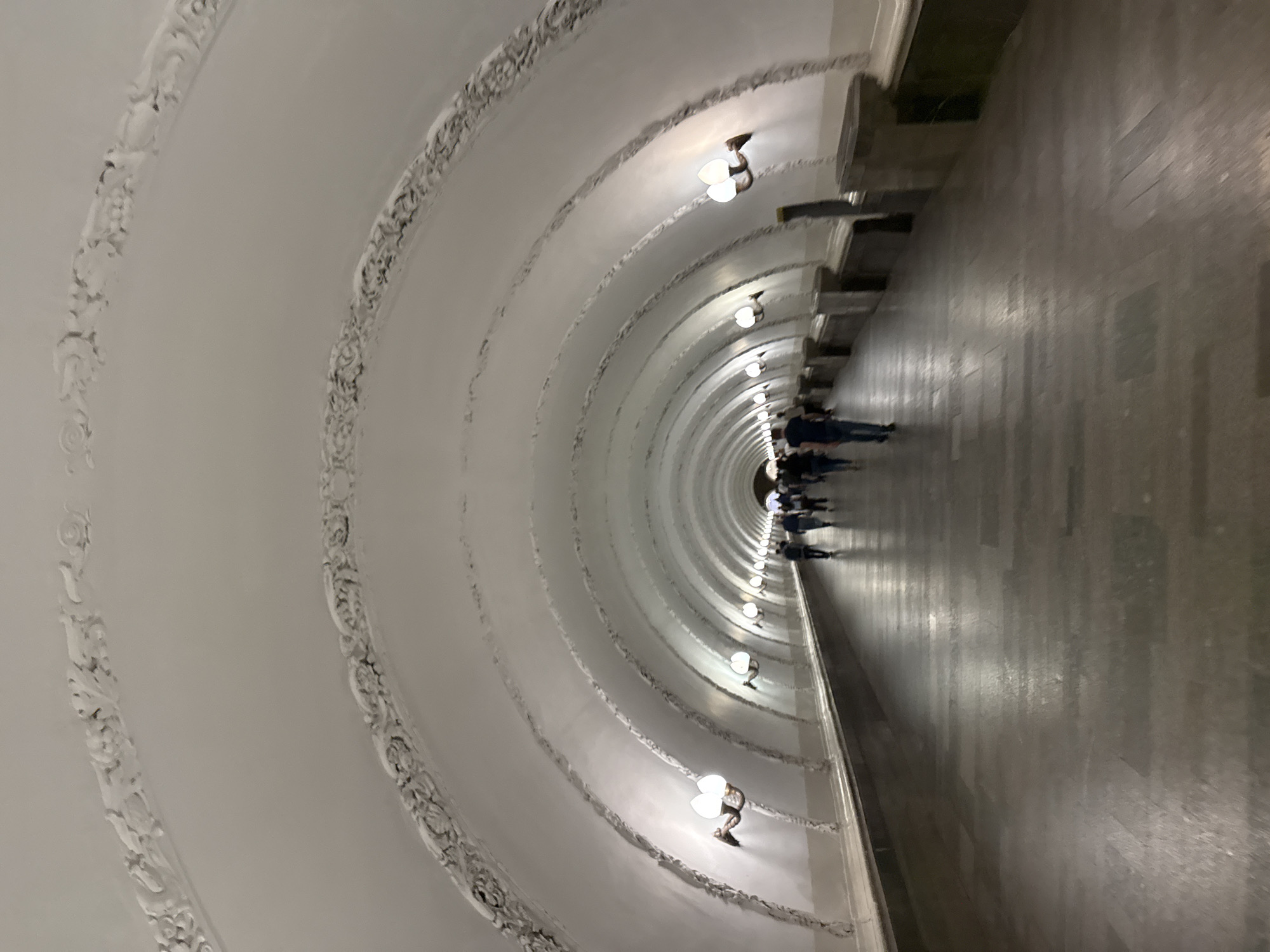 The image captures a long, ornate, deep tunnel or hallway, characteristic of the Moscow Metro, viewed from an angled perspective that makes the passage appear to curve upwards and to the left. The vaulted ceiling is a dominant feature, adorned with regularly spaced decorative white plasterwork or molding forming arches that recede into the distance. Each arch showcases intricate, scrolled patterns, some appearing darker with age or shadow, highlighting their elaborate design.
Illuminating the tunnel are numerous wall-mounted light fixtures, or sconces, with white, shell-shaped shades, casting a warm, yellowish glow along the curved surface. These lights are symmetrically arranged, following the curve of the tunnel and contributing to the sense of depth. On the right side of the image, a dark, polished floor, possibly made of stone or tile, reflects some of the artificial light. A continuous, dark ledge or base runs along the right wall, separating the floor from the ornate ceiling structure.
Several people are visible in the far distance, walking away from the viewer towards the vanishing point of the tunnel. Their forms are somewhat indistinct due to the distance and lighting, but they appear as small figures in dark clothing. The artificial lighting suggests an indoor setting, likely underground, meaning the time of day and weather are not relevant. The overall impression is one of grandeur and architectural beauty, commonly associated with historical metro stations. No visible text is present in the image.