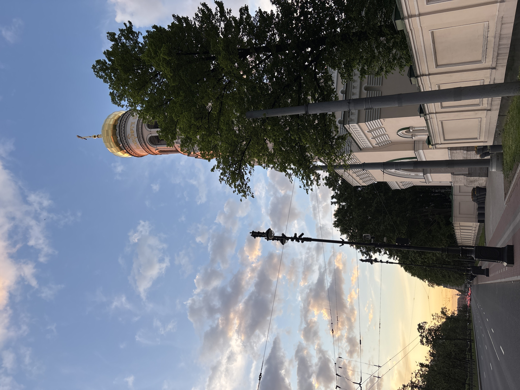 The image captures a quiet urban scene, likely an avenue in Saint Petersburg, Russia. A grand, light-colored building, which appears to be a church or cathedral, dominates the left side of the frame. It features classical architectural details, including pilasters and arched windows, and is crowned by a distinctive golden onion dome with a spire and cross. Cyrillic text is visible encircling the base of the dome, possibly a religious dedication.
Lush green trees line the street, partially obscuring the building and creating a natural canopy. A wide, multi-lane road stretches into the distance on the right, equipped with streetlights, utility poles, and overhead wires, suggesting trolleybus or tram lines. The sky is partly cloudy, displaying patches of clear blue interspersed with soft white and grey clouds. A warm, yellowish-orange glow on the distant horizon indicates either sunrise or sunset, bathing the scene in a tranquil light. No specific activity or large crowds are visible, conveying a sense of peace and quiet.