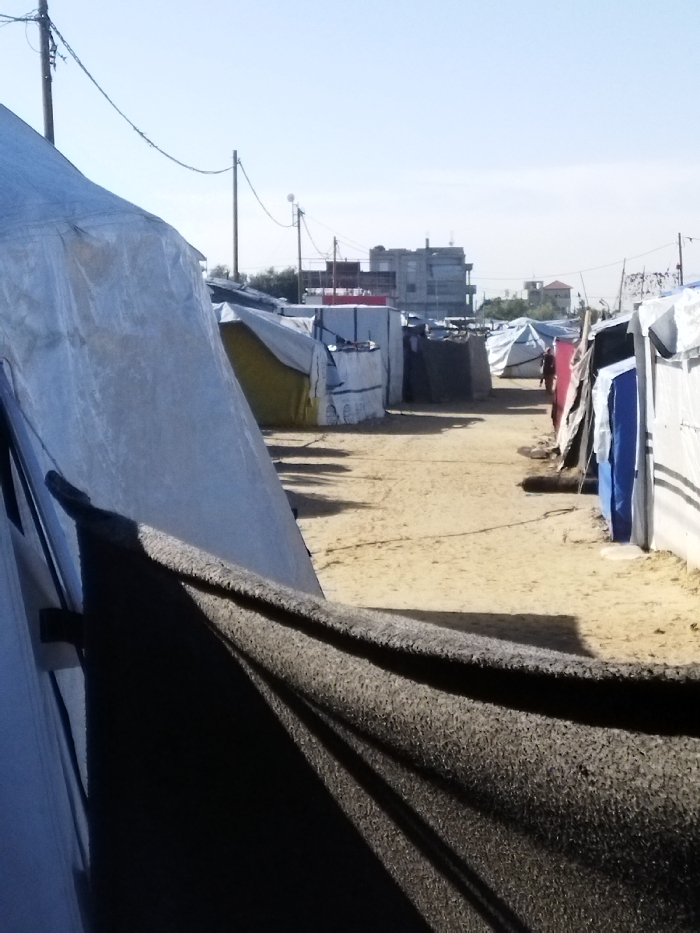 The image depicts a temporary settlement or displacement camp situated in Khan Yunis, Palestinian Territory, under a clear, light blue sky. A sandy dirt pathway, approximately 3-4 meters wide, recedes into the background, bordered by makeshift shelters on both sides. These shelters are constructed from various materials including light-colored tarpaulins (white, grey, yellow, blue, red) and corrugated metal sheets, forming low-profile, irregular structures. Shadows are cast onto the sandy ground from these structures. In the mid-distance, a lone individual in red attire walks away from the viewer on the right side of the path. Utility poles with overhead wires traverse the scene; one pole on the far left shows barbed wire near its apex. The background features several multi-story urban buildings, including a prominent dark building with rooftop equipment and a smaller structure with a red-tiled roof, indicating the camp's proximity to a developed area. The immediate foreground on the left is dominated by a large, light-colored tarpaulin tent, partially covered by a dark, textured fabric.