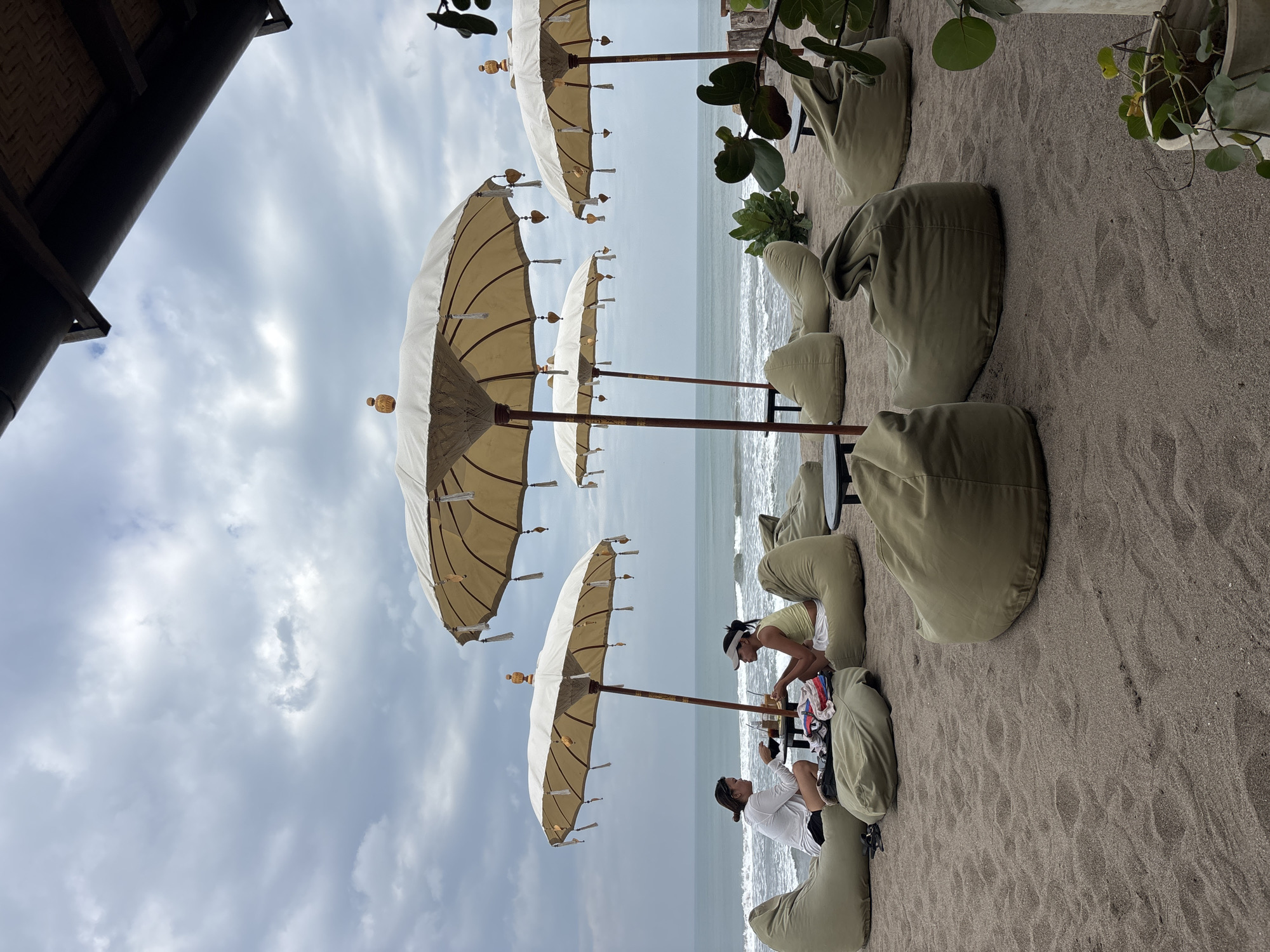 The image depicts a beachside seating area in North Kuta, Indonesia, characterized by several large, light-colored umbrellas providing shade over numerous olive-green beanbag chairs. In the background, the ocean waves gently roll onto a sandy beach under a cloudy sky. Two individuals are seated on beanbag chairs facing each other at a low table, engaged in conversation and holding drinks. The scene suggests a relaxed, casual atmosphere, likely a cafe or lounge situated on the coast.