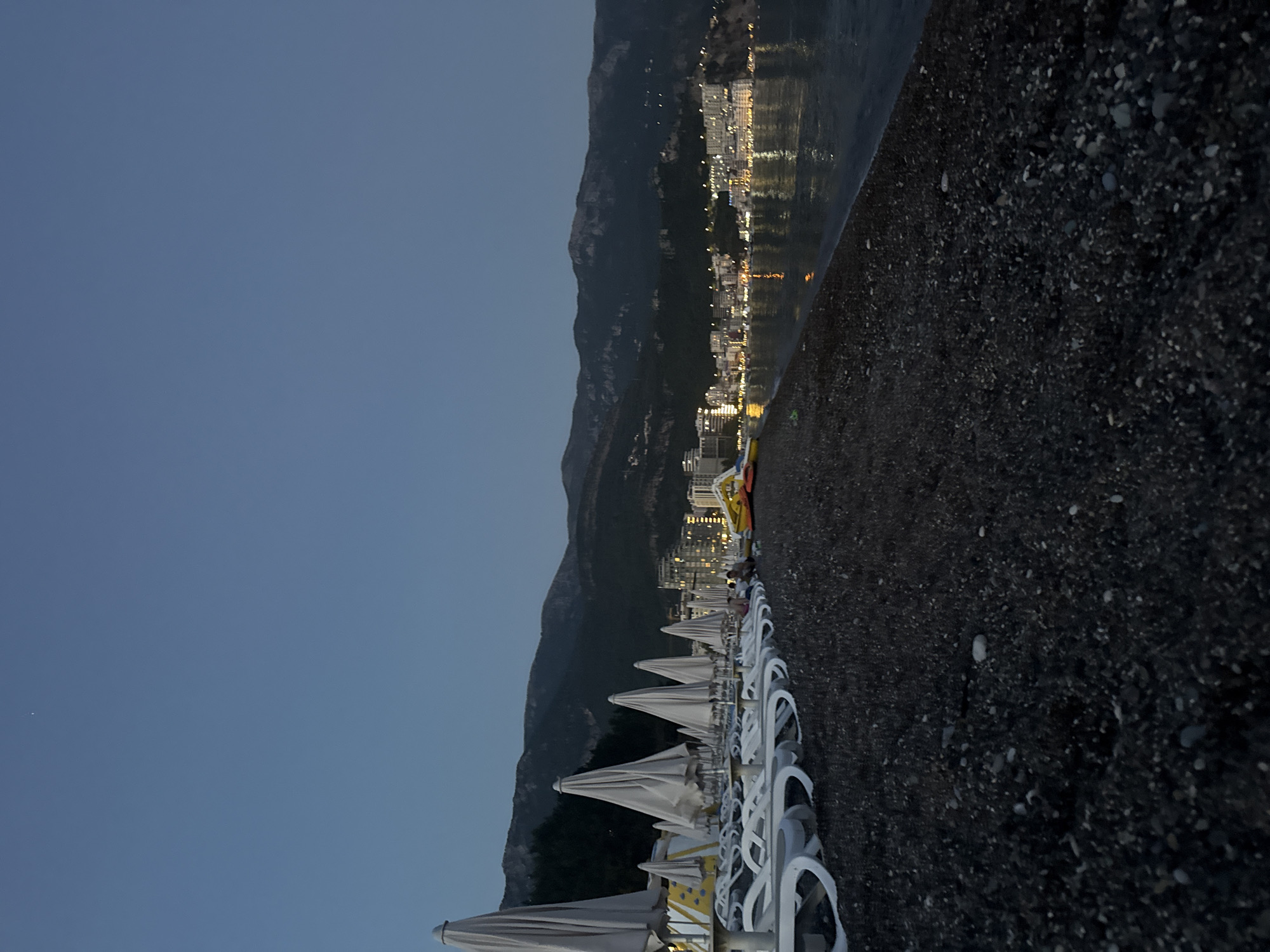 The image depicts a nighttime view of a beach in Boreti, Montenegro. A dark, pebble-strewn beach occupies the foreground, leading to the calm water's edge. A row of closed, white beach umbrellas and empty lounge chairs stretches across the middle ground. In the background, a coastal town is illuminated with numerous lights, nestled at the base of dark, mountainous terrain. The sky is a deep, clear blue, with a single bright star visible. The overall scene conveys a sense of quiet tranquility at dusk or after sunset.