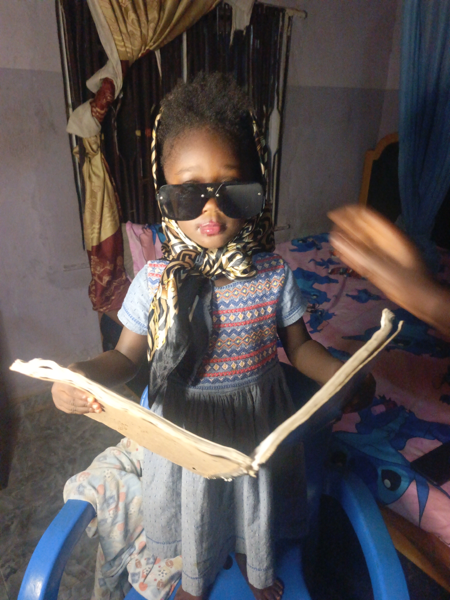 A young child stands on a blue plastic chair in what appears to be an indoor setting in Akwanga, Nigeria. The child is wearing dark sunglasses and a patterned headscarf, and is holding open a book with pale pages. The child is also wearing a blue dress with a patterned bodice and gray skirt. Behind the child, there is a curtained window. A blurred adult hand is visible on the right side of the image. A bed with a blue Stitch-themed blanket can be seen in the background.