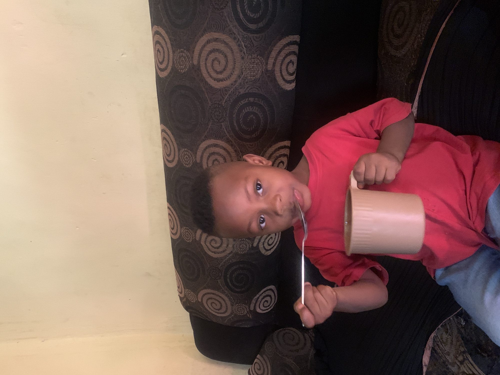 A young Black child, likely a boy, is pictured indoors in an informal setting. He is seated on a dark sofa or armchair upholstered with a distinctive pattern of brown and tan swirls and concentric circles. The child is wearing a bright red short-sleeved t-shirt and light blue pants.
In his right hand, he holds a light brown, possibly ceramic, mug with a handle. His left hand holds a silver utensil, appearing to be a spoon or fork, which is placed in his mouth. The child looks directly at the camera with a curious and engaged expression. The background to his left is a plain, light-colored wall, possibly off-white or light yellow. The overall impression is a candid moment, perhaps during a meal or playtime, captured indoors in Jos, Nigeria. No visible text is present.