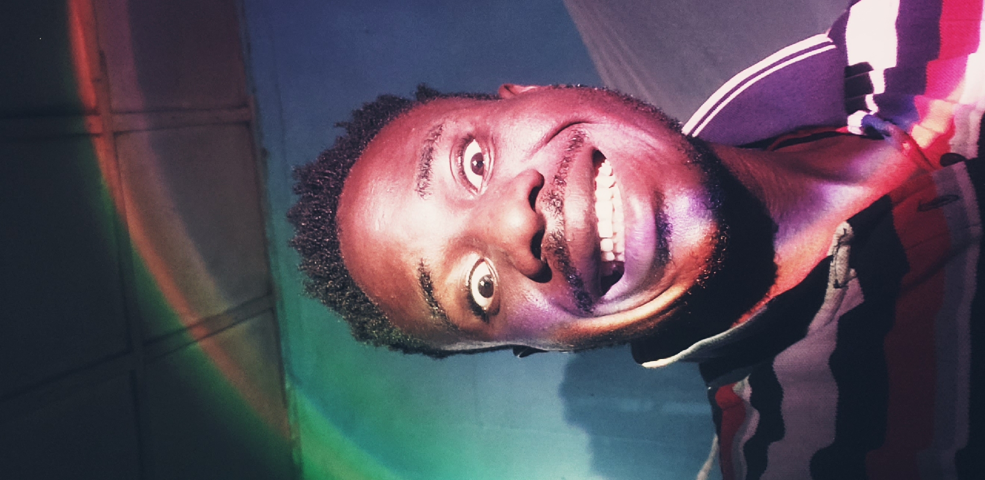 The image is a close-up selfie of a Black man with dark, short, curly hair and a beard. He is making a wide, exaggerated smile with his eyes wide open, giving him a surprised or playful expression. The man is wearing a collared shirt with horizontal stripes of black, red, and white. The background appears to be an indoor setting with a textured ceiling and a wall. There is a colorful light effect, resembling a rainbow or prism, cast across the upper left portion of the image and onto the man's face and the wall behind him. The lighting creates strong shadows and highlights, particularly on his skin, which has a reddish-purple tint in some areas due to the colored light. The scene is not explicitly identifiable as being in Narayi, Nigeria, based solely on the visual information provided.