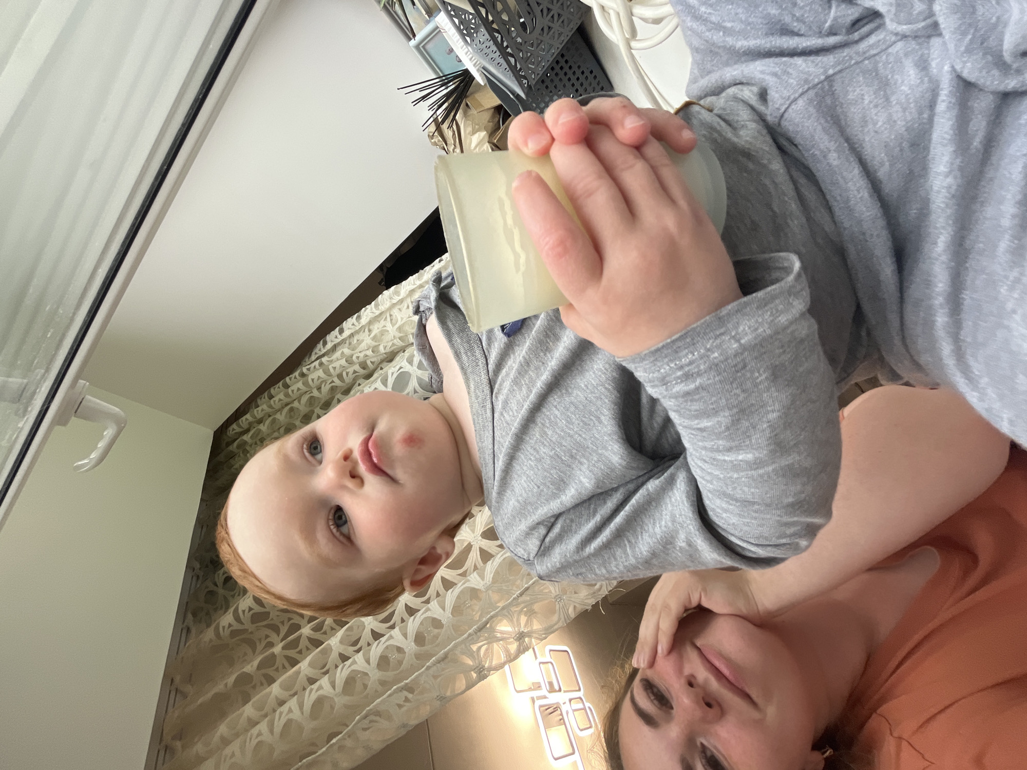 The media file shows two people, a young child and an adult, in an indoor setting. The child, with fair skin and red hair, is looking up with blue eyes and has a small red mark on their cheek. They are wearing a gray long-sleeved shirt and holding a translucent, light-colored object, possibly a cup or a candle. The adult, with dark hair and eyebrows, is also wearing a gray long-sleeved garment, and their arm is extended, with their hand resting on their forehead. The background features a patterned curtain and a white surface, possibly a table or a shelf, with some decorative items, including a gray woven basket containing what appear to be black sticks. A handle is visible on the left side of the image, suggesting it might be a door or window. The lighting appears to be indoor ambient light, and there is a reflective surface in the lower part of the image. The overall tone seems to be casual and intimate. The photo was taken in Chaikovsky, Russia.