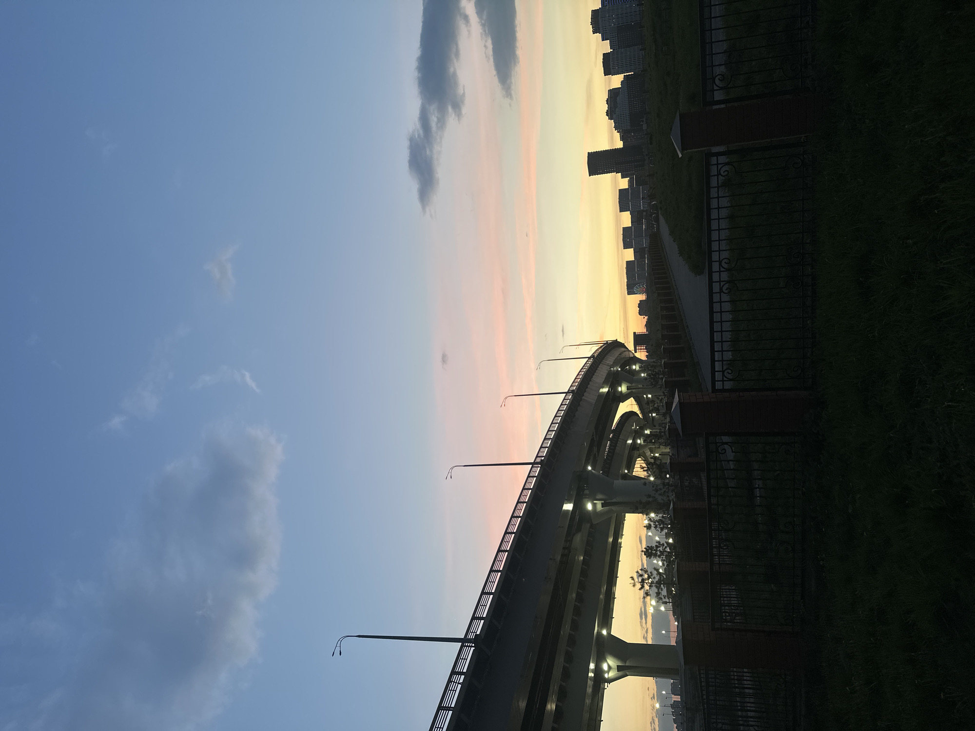 The image depicts an elevated highway in Kazan, Russia at sunset. The sky transitions from blue to orange and pink, with clouds visible. The highway has multiple levels and is illuminated by streetlights. Alongside the road, there's a paved path with a metal fence and brick posts, bordering a grassy area. In the background, the silhouettes of tall buildings define the skyline.