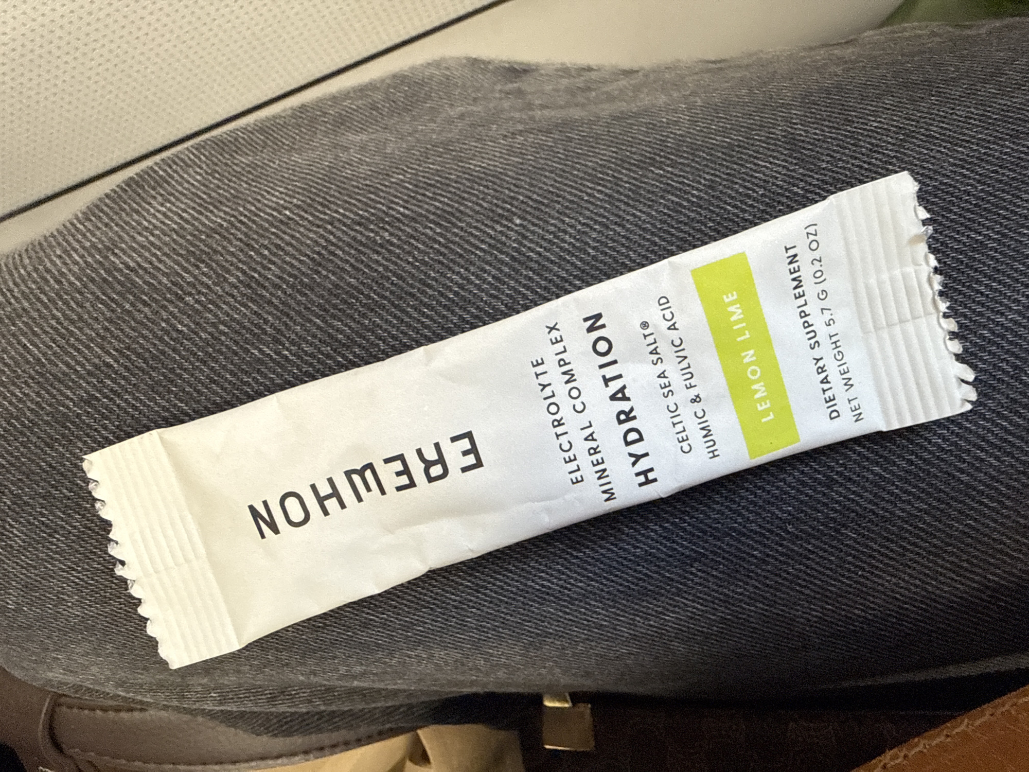 The image shows a white Erewhon hydration packet resting on what appears to be a pair of gray denim pants. The packet is labeled "ELECTROLYTE MINERAL COMPLEX HYDRATION" with a "LEMON LIME" flavor indicated on a green section. It also notes that it is a "DIETARY SUPPLEMENT" with a "NET WEIGHT 5.7 G (0.2 OZ)".
Erewhon is a chain of organic grocery stores located in Los Angeles, California.