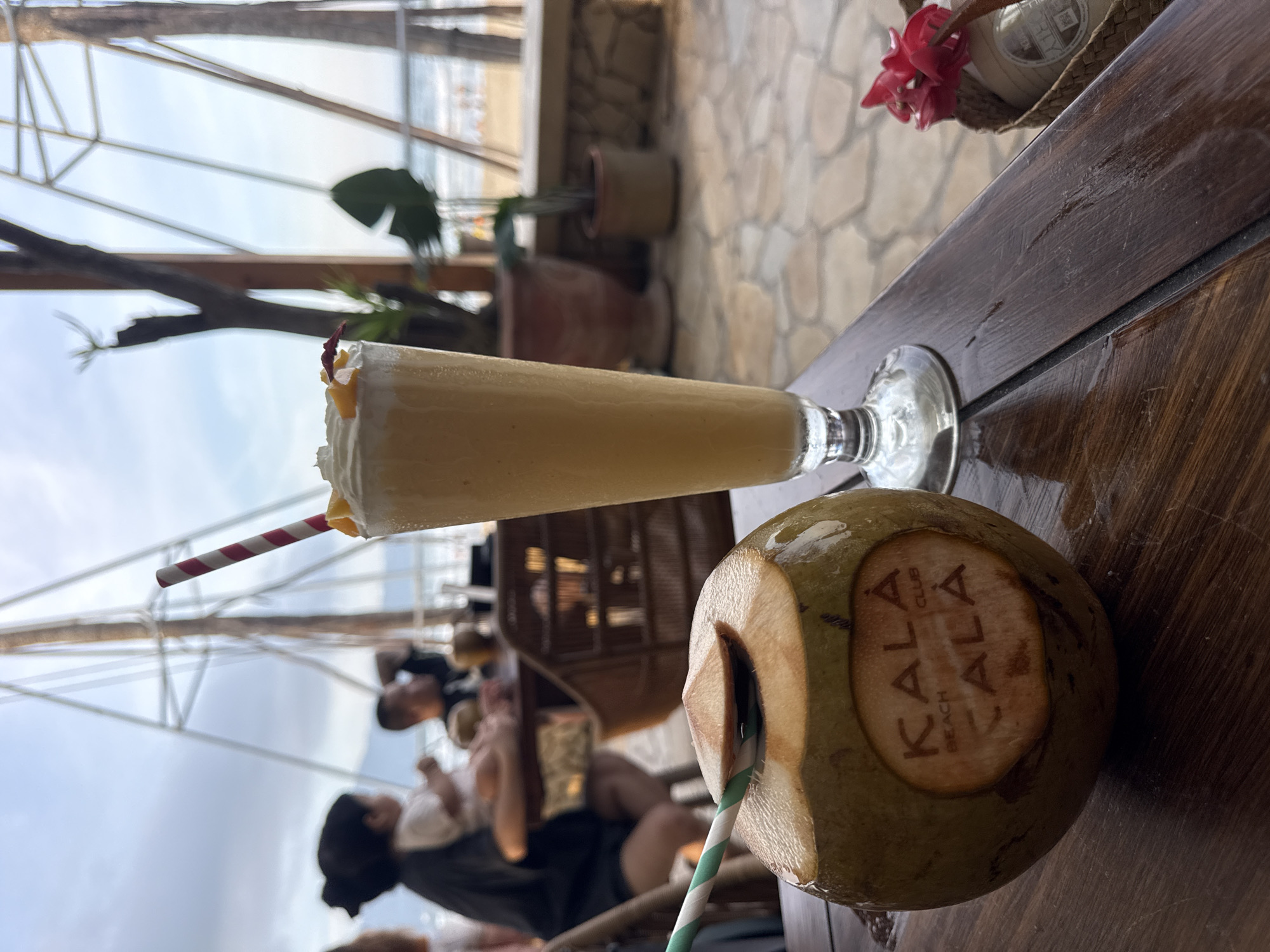 The image shows a tropical scene at the Kala Beach Club in an unknown city. The focus is on a wooden table with a coconut drink, featuring the "KALA BEACH CLUB" logo, and a milkshake in a glass, each with a straw. In the background, several indistinct people are sitting in wicker chairs, possibly patrons of the beach club. The background reveals a beach scene with an ocean view under a partially obscured sky. The building architecture includes wooden beams and a stone wall, suggesting a rustic, beach-themed setting.