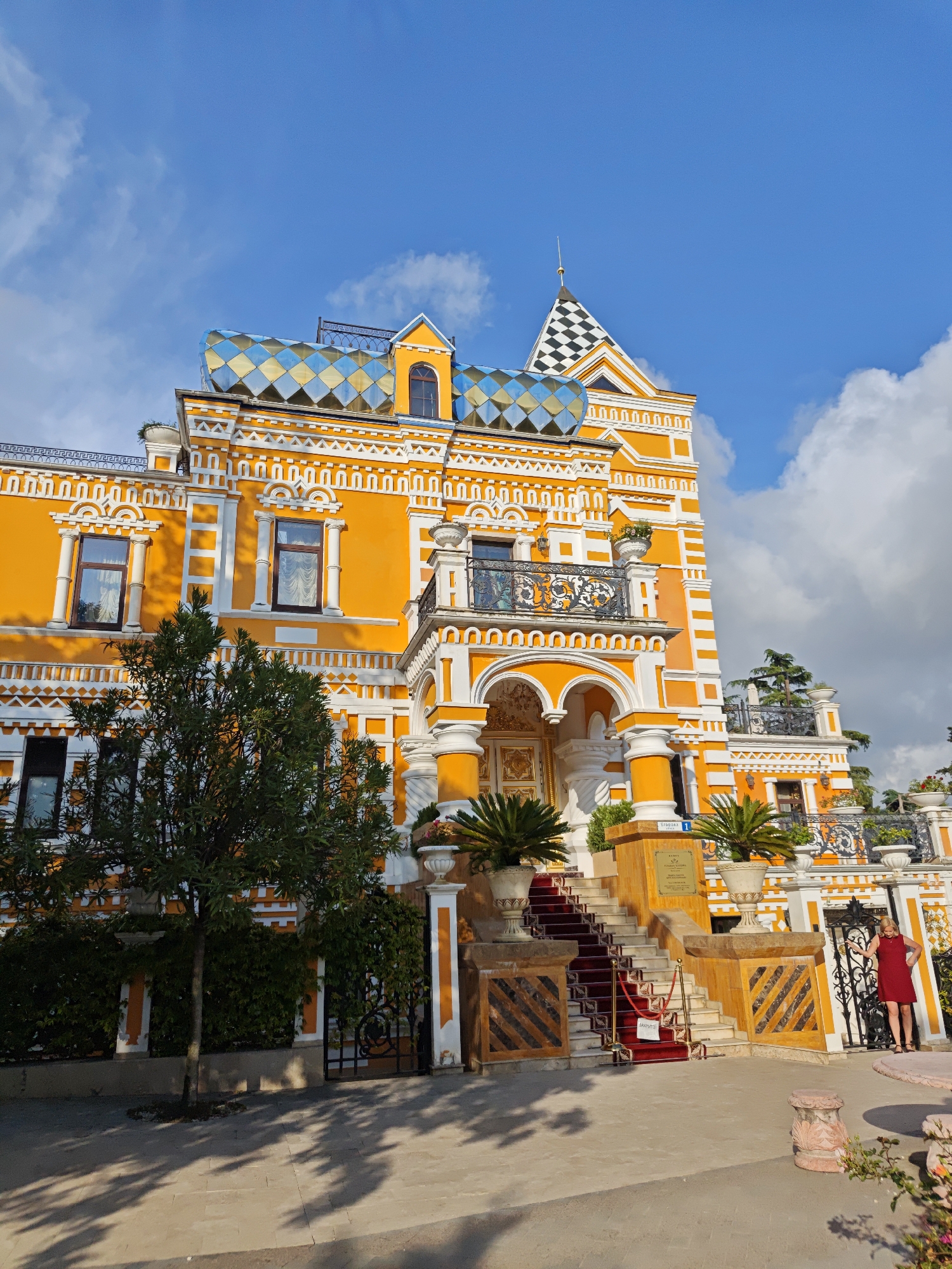 The image shows the exterior of the historic Villa Nadezhda in Sochi, Russia. The building is a striking three-story structure, primarily orange with white trim. Its architectural features include decorative window frames, an ornate balcony with a wrought-iron railing, and a unique roof with a blue-and-silver diamond pattern. A tower with a black-and-white checkered design also accents the roofline.
The entrance is marked by a set of stairs covered with a red carpet and flanked by decorative urns with plants. A tree partially obscures the left side of the building, and a wrought-iron gate is on the right. A woman in a red dress stands near the gate. The sky is blue with scattered clouds, indicating a clear day. The scene is well-lit, casting shadows on the ground.