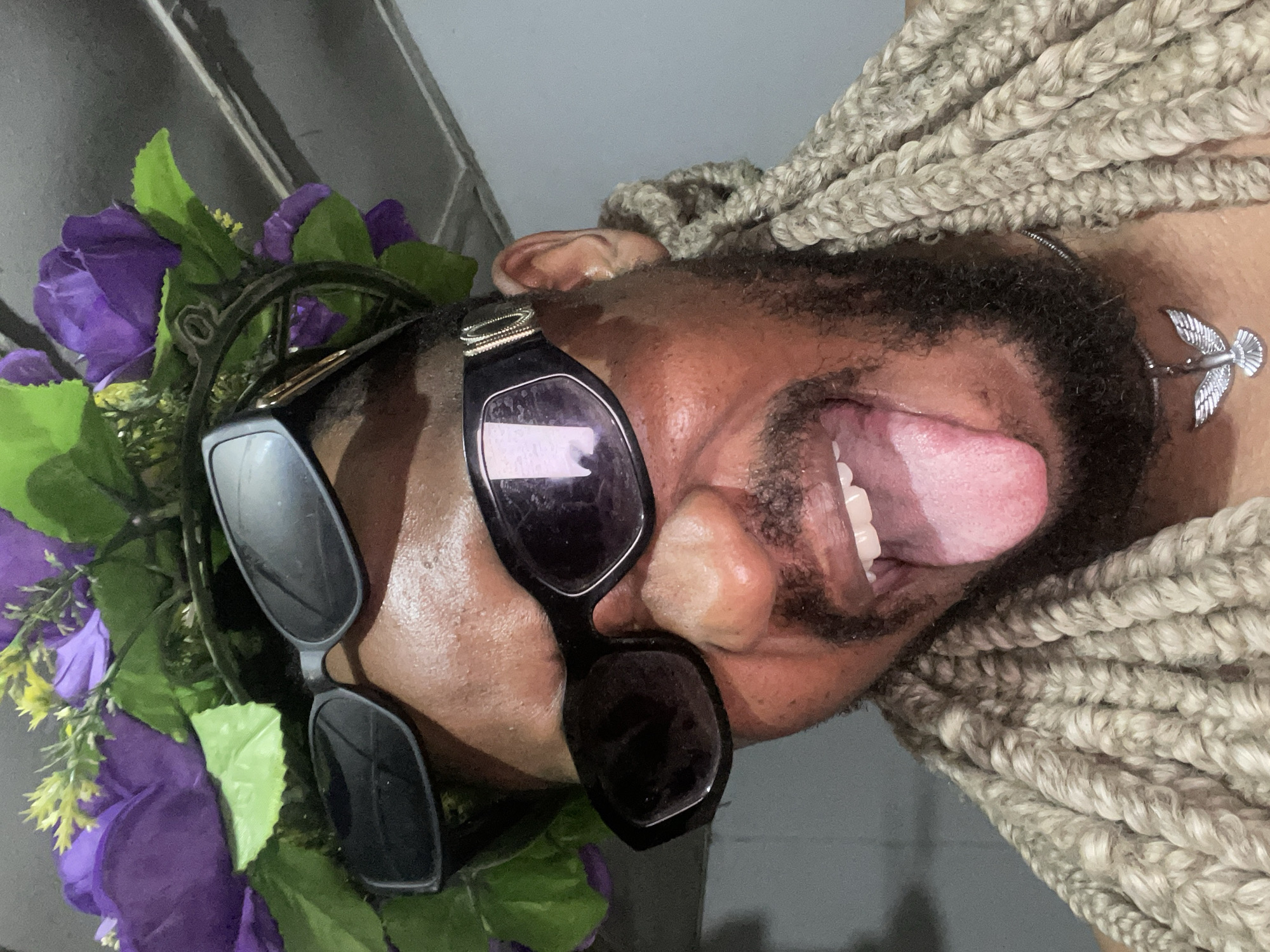 A close-up, tilted portrait captures a Black person wearing two pairs of sunglasses and a floral crown of purple flowers and green leaves. The individual has beige braids, a beard, and a silver bird-shaped necklace. Their mouth is open, revealing their tongue and teeth. The background is a blurred grey, and there is no indication of the location being Jos, Nigeria.