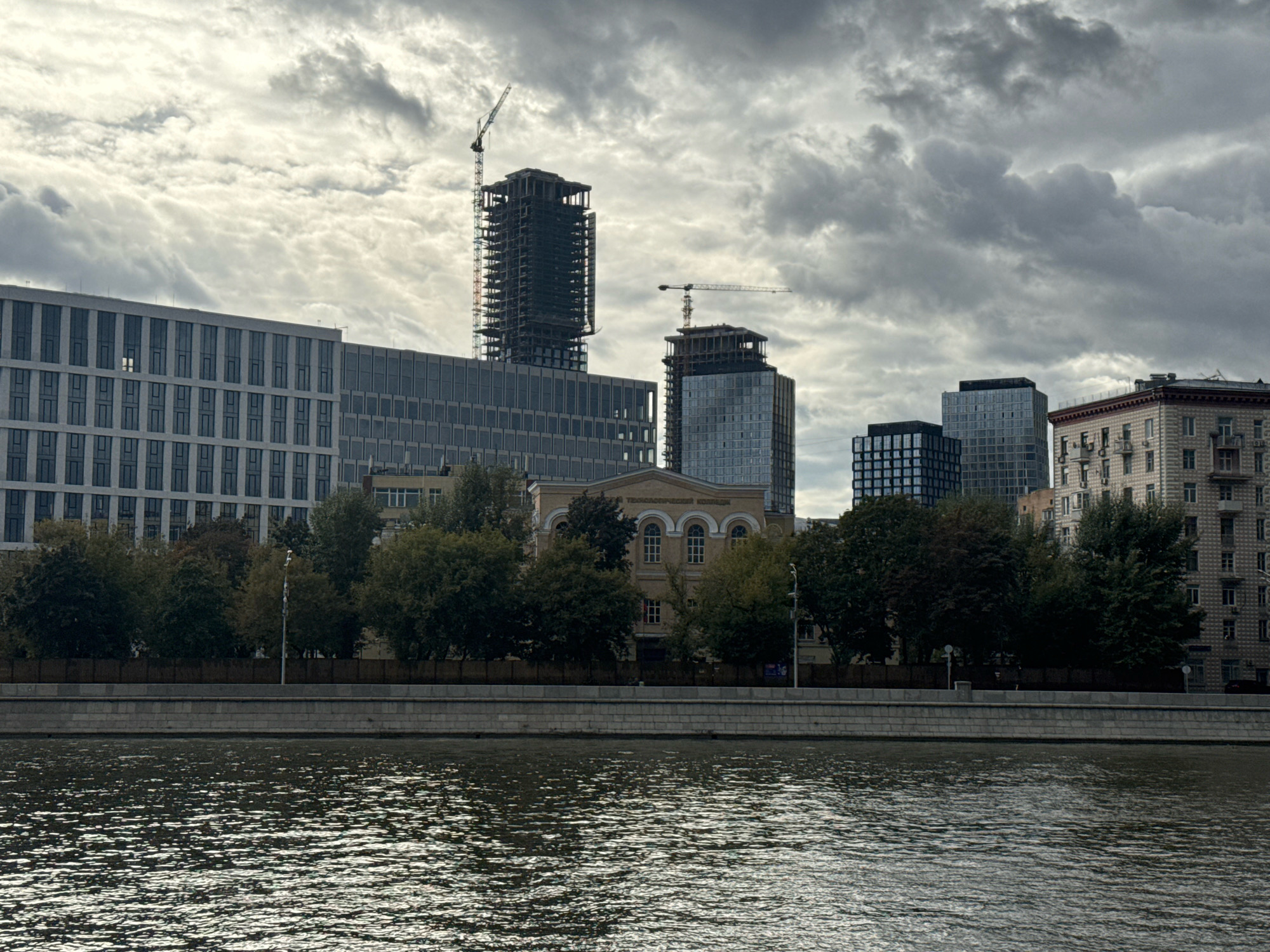 The image shows a body of water in the foreground, with a stone embankment behind it. Beyond the embankment, a row of trees lines the bank. In the background, a city skyline is visible, featuring a mix of modern skyscrapers, some of which are under construction with cranes, and older buildings. One of the older buildings appears to be a historic structure with arched windows and has Cyrillic text on its facade that reads "ТЕХНОЛОГИЧЕСКИЙ КОНТРОЛЬ", suggesting it is related to technological control or inspection. The sky is overcast with dramatic, dark clouds. The scene is likely in Moscow, Russia, given the presence of Cyrillic text and the architectural style of some of the buildings, potentially along the Moskva River. There are no visible people or animals in the image.