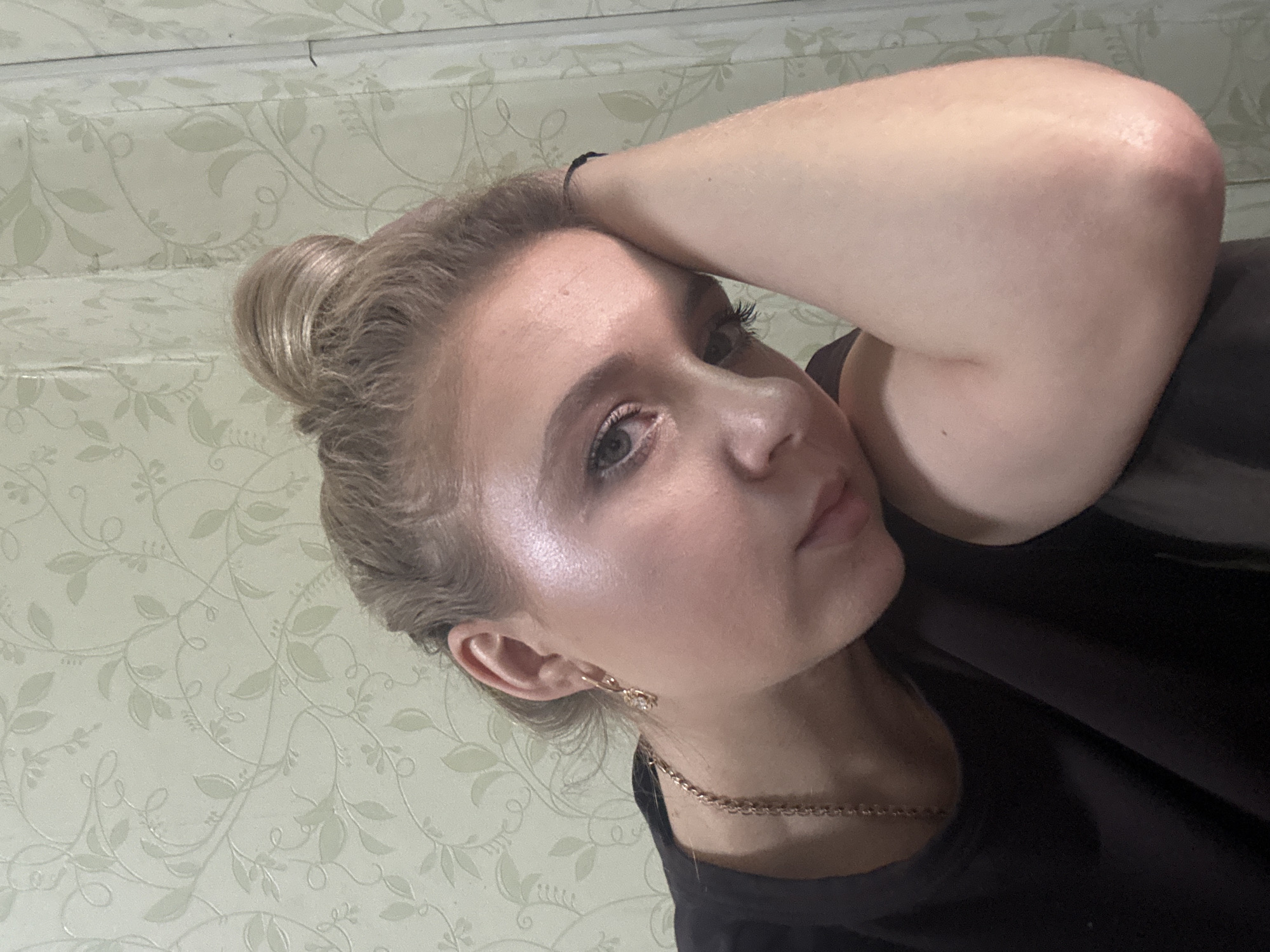 A young woman with blonde hair pulled into a top knot rests her head on her arm. She is wearing a dark t-shirt and a gold chain necklace. She has on an earring in her left ear. The background is a light green wallpaper with a floral pattern. She is looking directly at the camera.