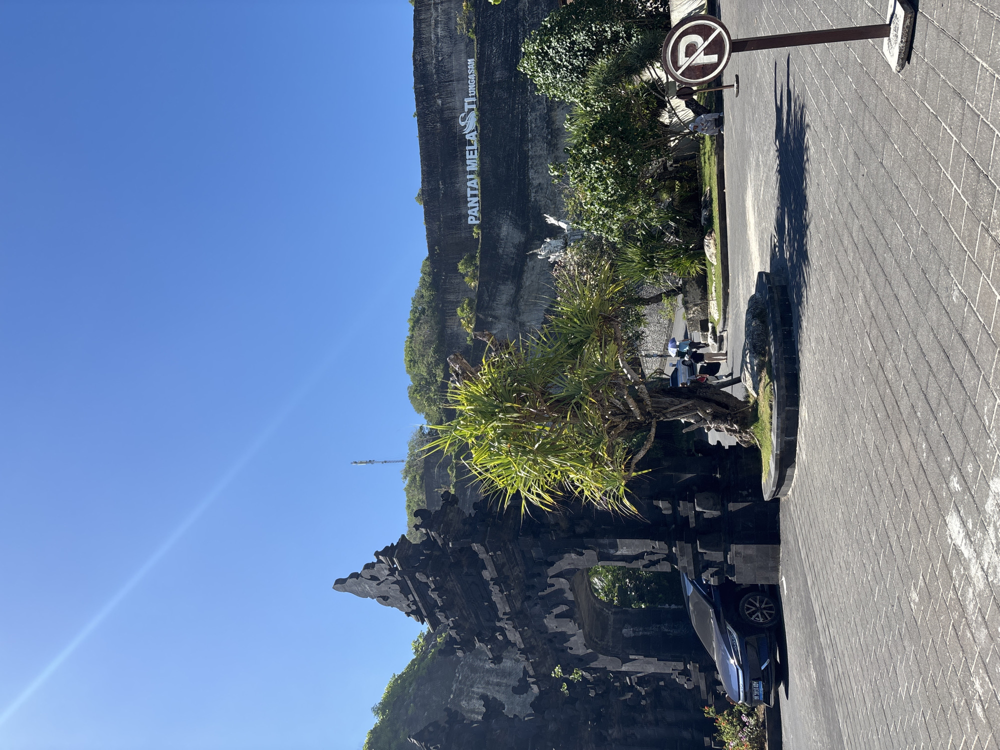 This image shows the outdoor parking area at Pantai Melasti Ungasan in Kuta Selatan, Indonesia. The sky is clear blue. The parking area is paved with interlocking grey stones. A 'no parking' sign stands next to a small area with a tree. A blue car is parked on the other side of this area. In the background, a large cliff face features the words "PANTAI MELASTI UNGASAN" written in large white letters. Green trees and vegetation surround the cliff face.