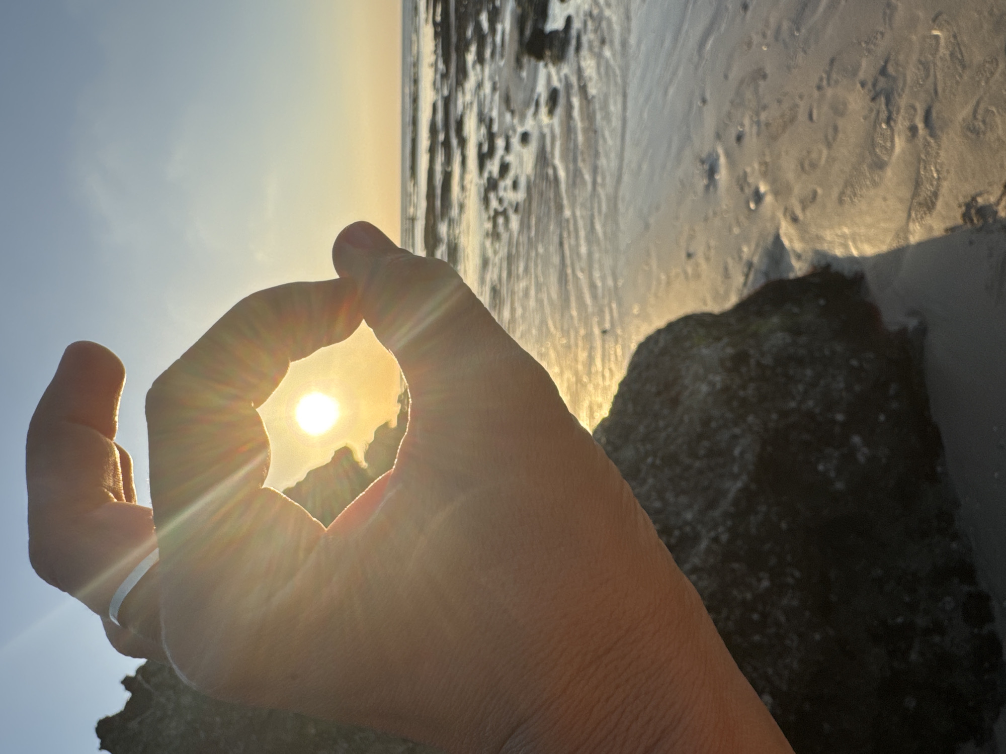 A hand forms an "O" shape, framing the setting sun against a clear sky. The sun's rays emanate outwards, creating a lens flare effect. The hand, with a ring on the index finger, is positioned in the foreground, obscuring a portion of a rock. In the background, a beach stretches towards the ocean, with calm water and scattered rocks. The sand is wet, reflecting the warm light of the sun. The scene appears to be a coastal landscape during golden hour. The location is Kuta Selatan, Indonesia.