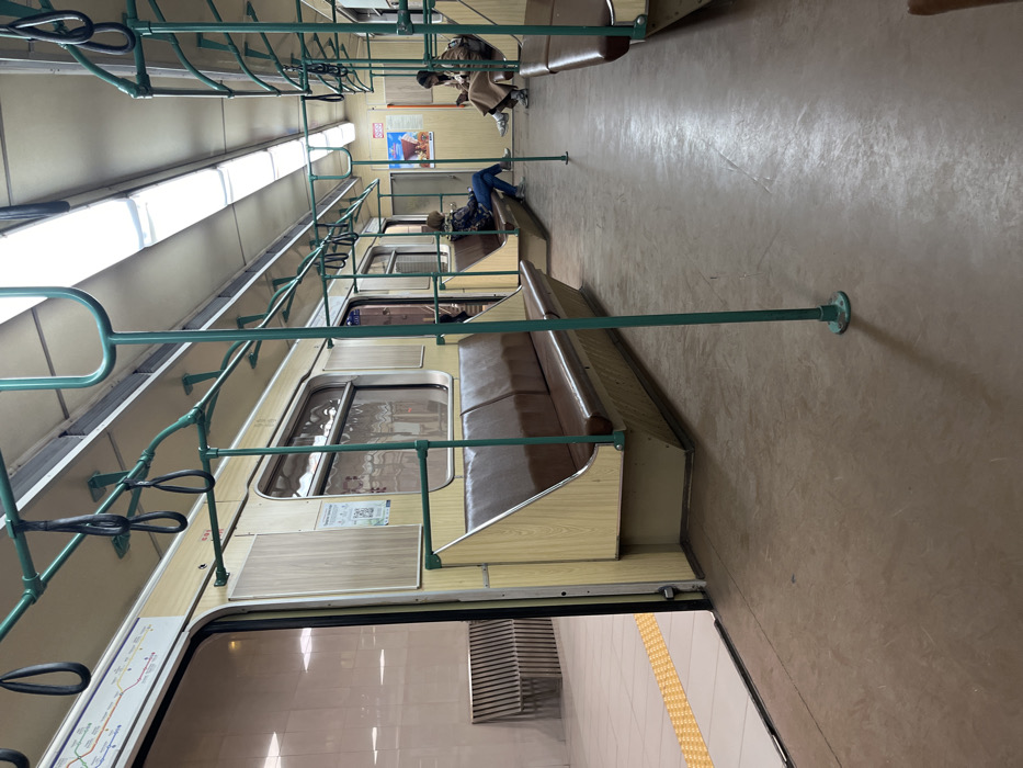 The image displays the interior of a Sofia Metro train car, currently stationary with its doors open to a platform in Sofia, Bulgaria. The train car features light wood-paneled walls and seat bases, brown cushioned seating, and a system of green metal handrails and vertical poles from which black looped hand straps are suspended overhead. The floor is brown, and two elongated fluorescent light fixtures illuminate the beige ceiling.
Three individuals are present inside the train car. On the left side of the aisle, a person wearing a checkered top and blue jeans is seated, looking downwards. Further down the right side of the aisle, another person dressed in a light-colored coat is seated, observing a small white device in their hands. A third individual is partially visible, seated behind the second person on the right.
Through the open doors on the left, a subway platform is discernible. It features white tiled flooring, a yellow tactile warning strip along the edge, and a slatted wooden bench against the platform wall. A multi-colored metro map is affixed to the train wall above the open door, and a small QR code sticker is visible on an adjacent wall panel.