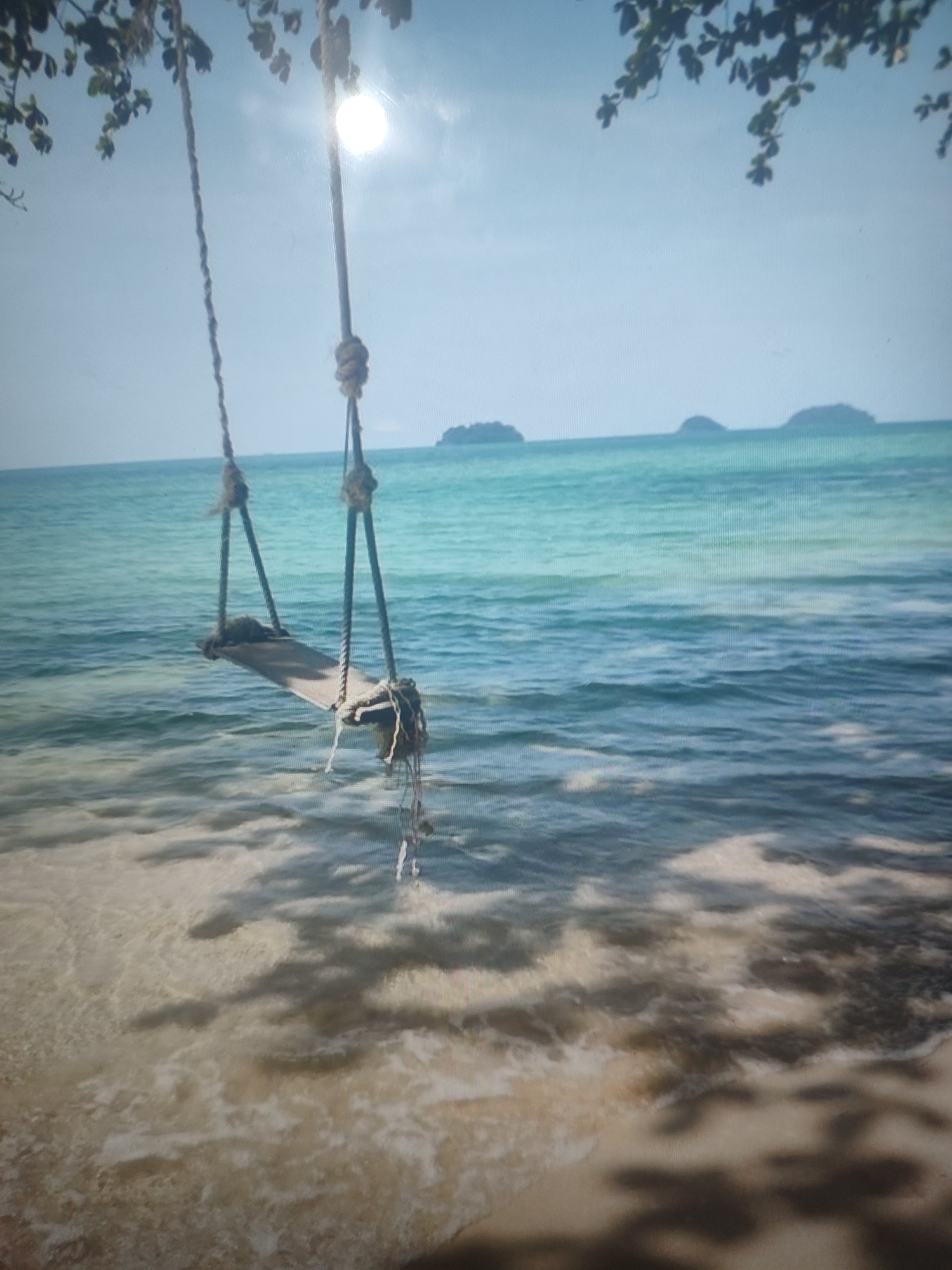 The image shows a swing hanging over clear blue water on a beach. The swing is made of rope and a wooden plank seat. The sun is shining brightly, and there are islands in the distance. The water is calm and reflects the sky. The beach is sandy and has shadows from trees. There is no indication of the location being in Chernivtsi, Ukraine.