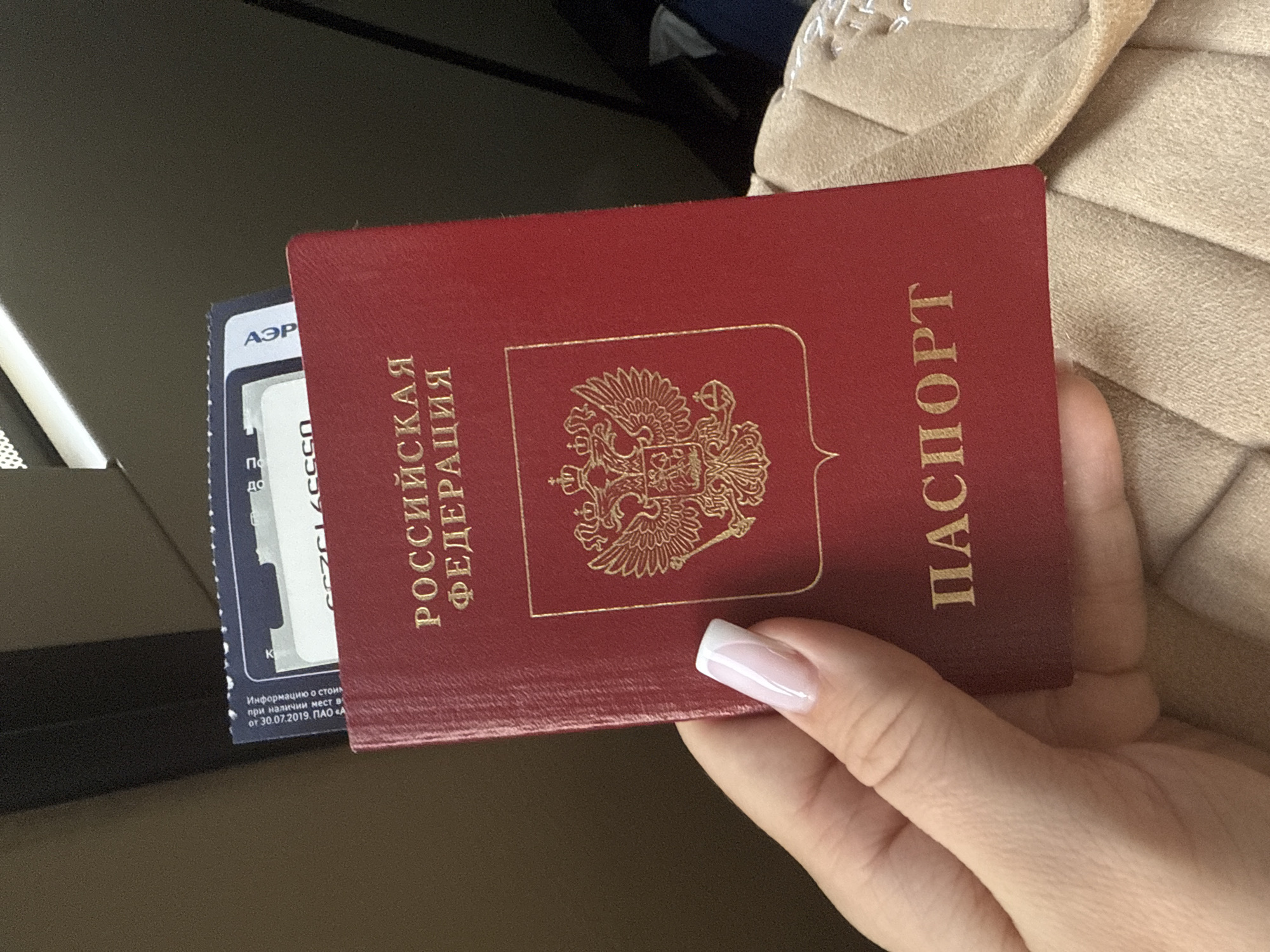 A hand with a French-tip manicure holds a Russian passport and a blue boarding pass from Aeroflot airline, with text that includes a date of July 30, 2019. The passport is red with the Russian coat of arms and the words "Russian Federation" and "Passport" in gold lettering. The location in the city of Khimki, Russia, cannot be definitively determined from the image.