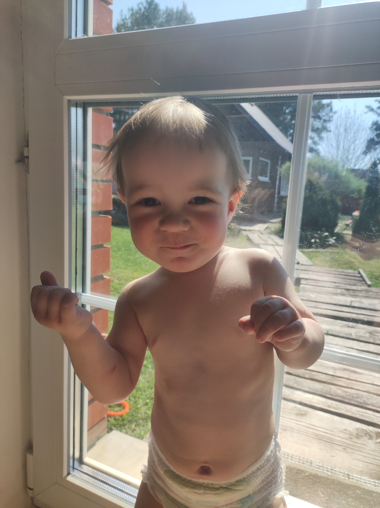 A toddler is standing in front of a window. The toddler is fair-skinned, with blondish hair, and wearing a diaper. The toddler is looking directly at the camera with a slight smile. The window has multiple panes and appears to be overlooking a backyard or garden area. Trees, grass, and a building are visible through the window. The sun is shining brightly, illuminating the scene.