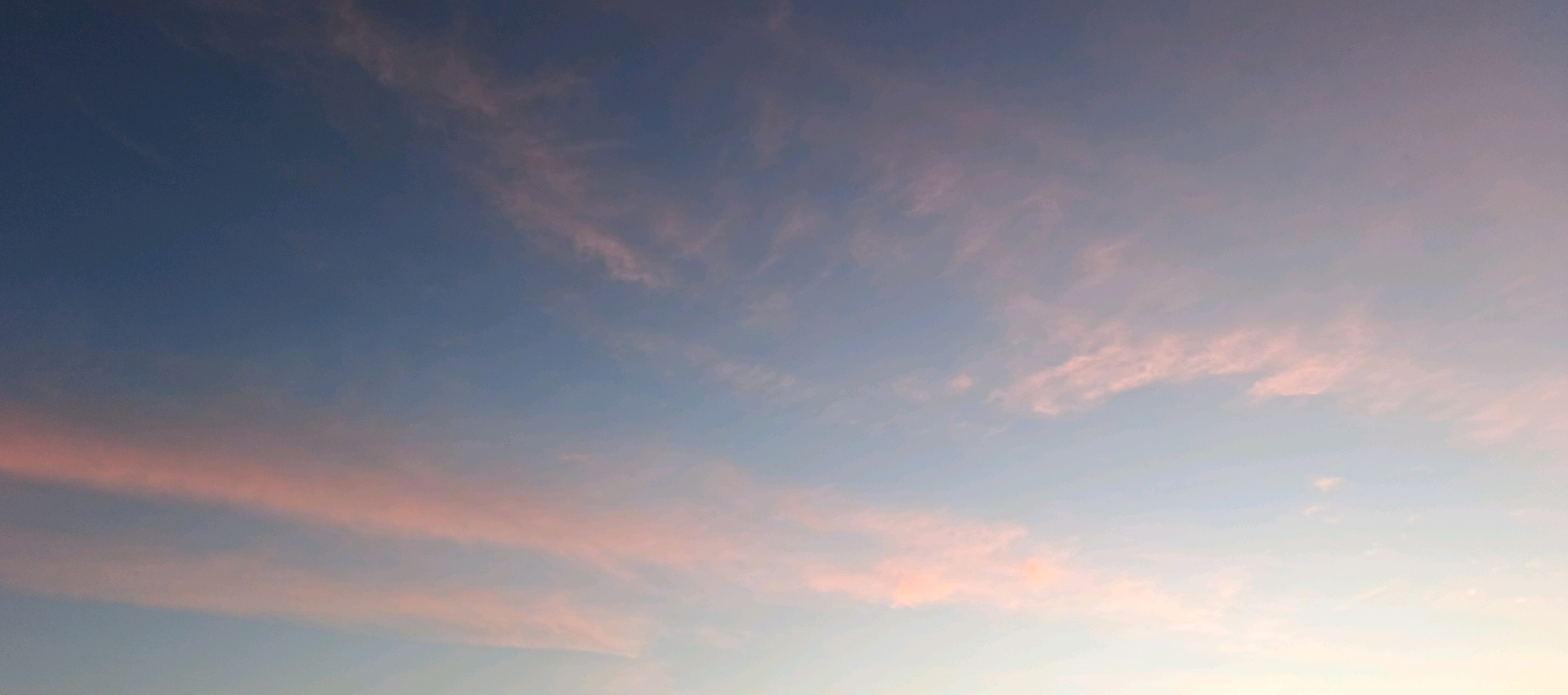 The image depicts a serene sky over Torbeevo, Russia, characterized by a gradient of colors transitioning from deep blue at the top to lighter hues of pink and white towards the horizon. Wispy clouds are scattered across the sky, tinged with soft pink from the setting or rising sun. The scene is entirely natural, with no human presence or man-made structures visible. The environment is solely the sky, showcasing the atmospheric conditions and colors of twilight.