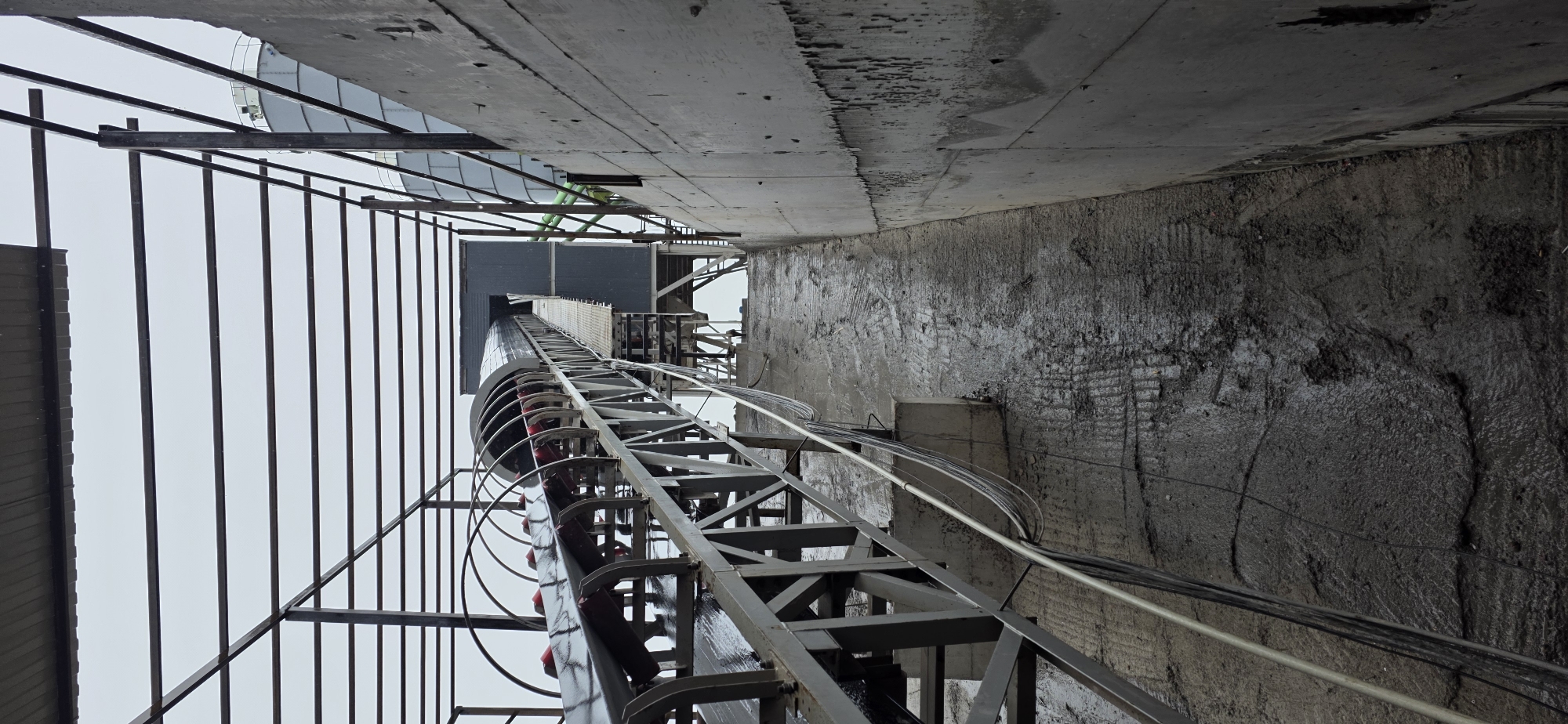 The image shows a view of an industrial site in Jyrgalan, Kyrgyzstan, under an overcast sky. A conveyor belt system, supported by a metal truss structure, ascends from the foreground towards a building or structure in the background. The conveyor belt itself is covered with a curved housing and appears to be wet, as does the concrete ramp or walkway leading up to it, suggesting recent rain or wet conditions. Cables run along the side of the conveyor structure and down the ramp. To the right of the ramp is a tall, textured concrete wall. In the far background, two large cylindrical silos are visible, typical of industrial facilities. The overall scene depicts an active or recently active industrial area with functional machinery.