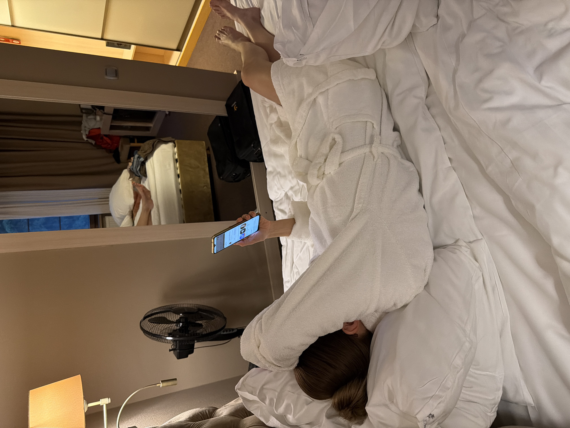 An overhead-angle image captures a person lying prone on a white bed in what appears to be a hotel room in Svetlogorsk, Russia. The individual, possessing light brown hair styled in a bun, has their head turned to the left, resting on a white pillow, with their left ear visible. They are clad in a white terrycloth bathrobe, which covers their upper body and is partially draped over their head. The right arm is extended, holding a gold-framed smartphone with an illuminated screen displaying a user interface featuring multiple circular profile images and text, consistent with a social media or messaging application. The bed is furnished with white sheets and a white duvet.
To the left of the bed, a bedside lamp with a conical, amber-colored fabric shade emits a warm light, positioned above a black oscillating fan. A large mirror dominates the wall opposite the bed, reflecting elements of the room. In the mirror's reflection, bare feet are visible, resting on a white duvet of an adjacent or the same bed, implying either another person or the subject's own legs from a different angle. Below these reflected feet, two black suitcases are discernible on the carpeted floor. Further into the reflection, a window draped with dark brown curtains and a bedside table laden with clothing and a red bag are visible. To the far right of the primary image frame, a segment of a light-colored wardrobe with sliding doors is present. The room's ambient lighting is low, supplemented by the bedside lamp and the phone screen.