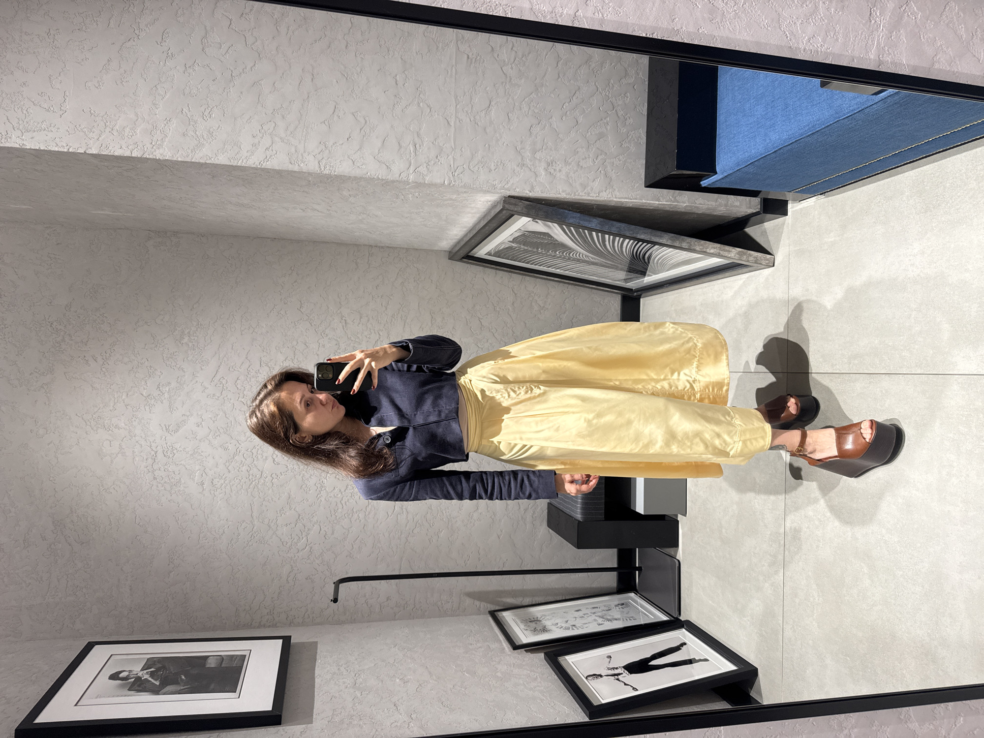 The image shows a woman taking a selfie in a room that appears to be a clothing store or showroom. She is wearing a dark blue jacket, a yellow skirt, and brown platform sandals. She is holding a black smartphone.
The room has a modern, minimalist design. The walls are light gray with a textured finish. There are several framed black and white photographs on the walls and leaning on the floor. A dark-colored clothing rack with a horizontal bar is visible. Above, a blue upholstered bench or seat is partially visible.
The overall impression is that the woman is trying on clothes in a shop. It is not possible to discern the specific location within Dubai, United Arab Emirates, based on the visual information provided.