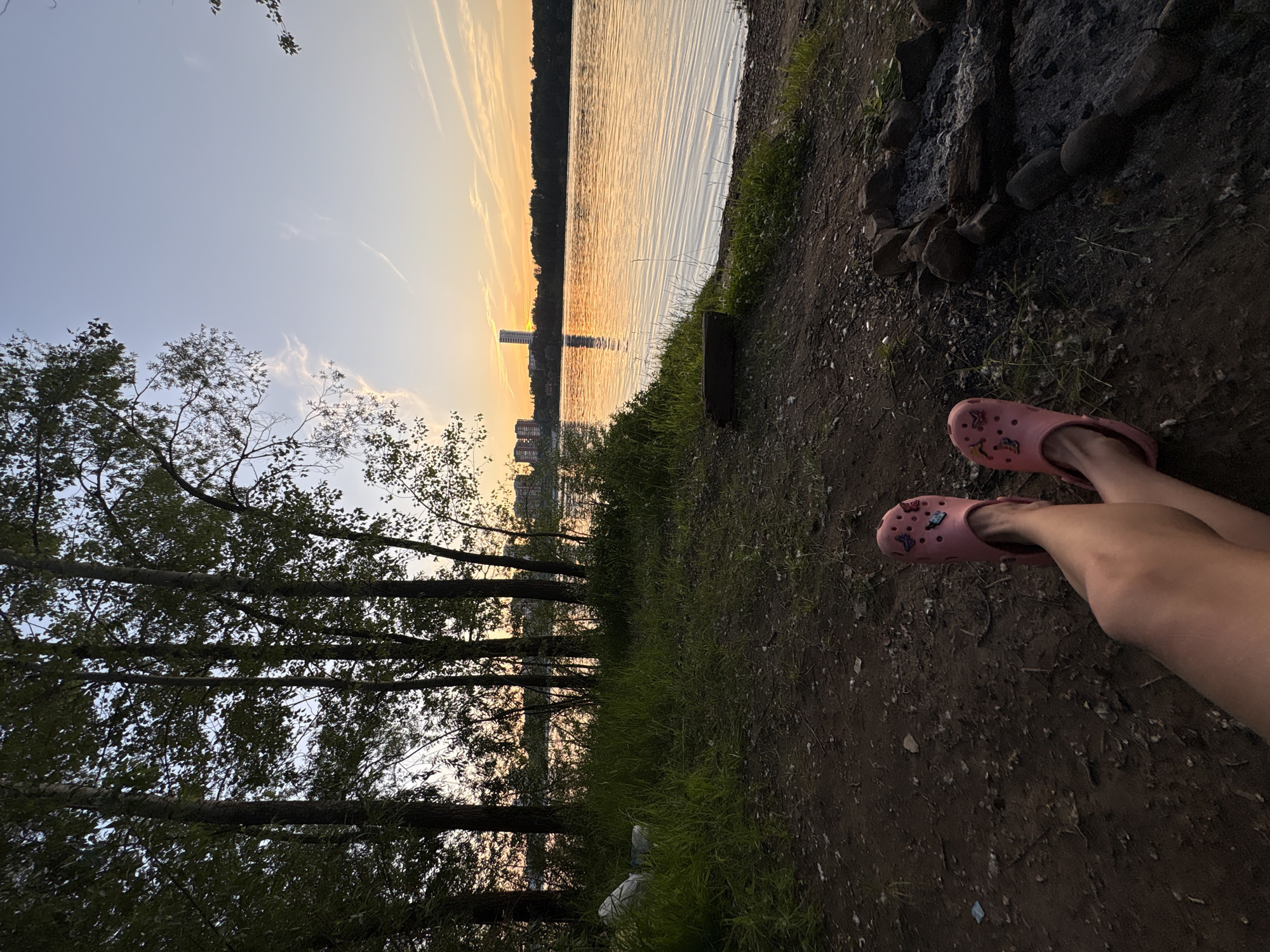 A person is sitting on the ground by a lake during sunset, with their legs crossed and wearing pink Crocs. The lake reflects the orange sky, and trees line the shore. In the distance, buildings are visible. A fire pit is located to the right of the person. The scene appears to be in a natural area within or near the city of Perm, Russia, possibly along the Kama River.