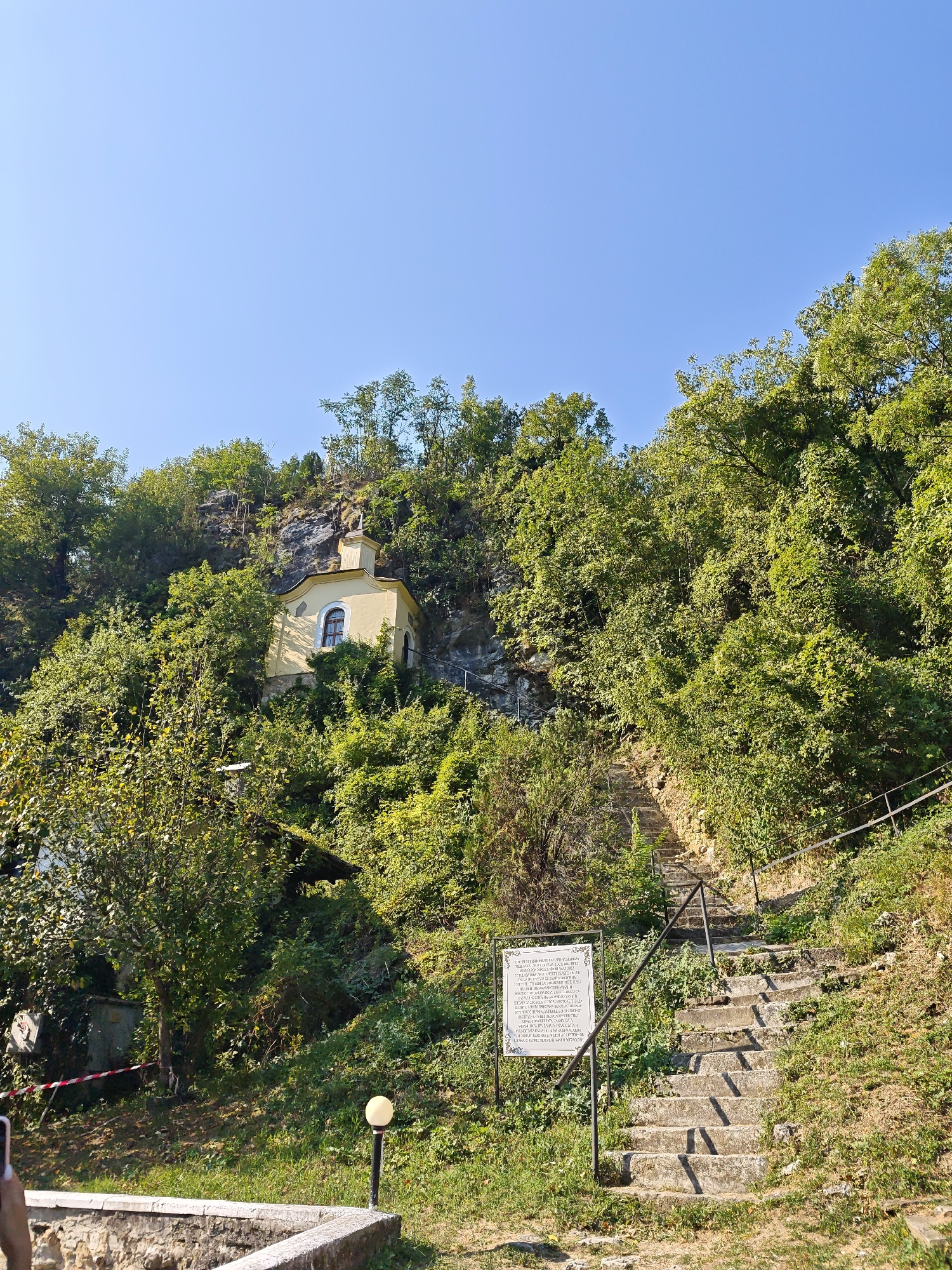 The image shows a small, yellow chapel built into a rocky hillside, surrounded by lush green trees. A stone staircase with a metal railing leads up the hill towards the chapel. A white sign with text is posted near the bottom of the stairs. The sky is clear and blue. The scene appears to be the Chapel of Saint George near the village of Lyutibrod, Bulgaria.