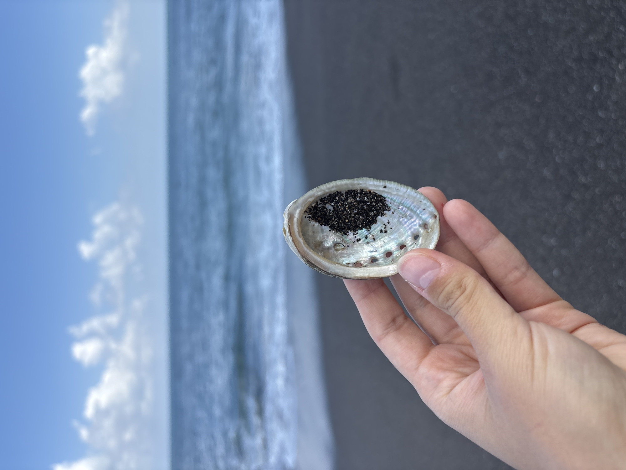 A hand holds a seashell filled with black sand against a blurred background of a beach and ocean. The seashell is iridescent, showing pearly white and bluish-green colors. The black sand is piled inside the shell, with some grains scattered on the inner surface. The ocean water is a deep blue, with small waves breaking on the shore. The sky above is a clear blue with scattered white clouds. The image appears to be taken on a sunny day. The location is identified as Mengwi, Indonesia.