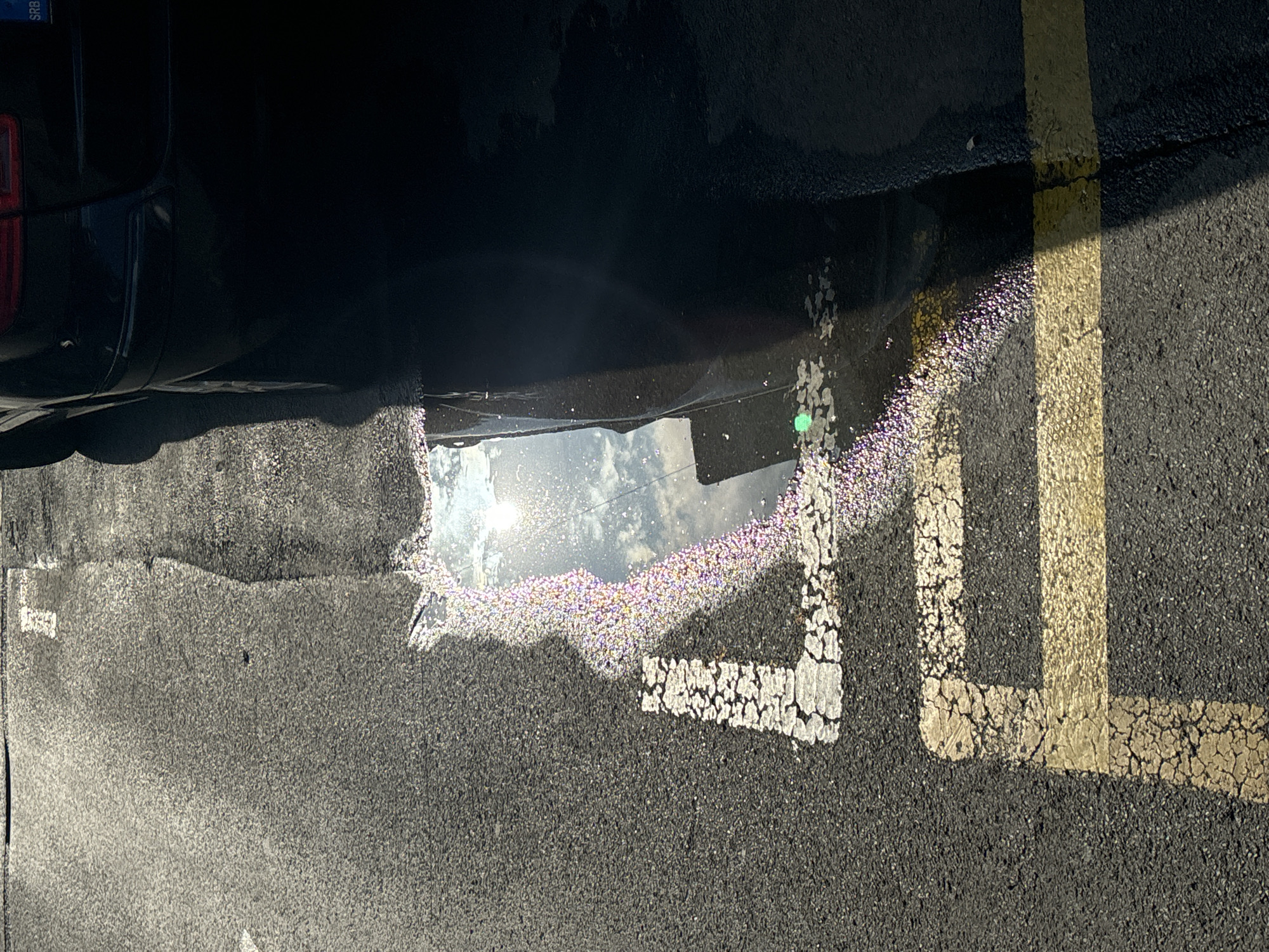 In Belgrade, Serbia, a puddle reflects the sun and sky on asphalt marked with faded yellow and white parking lines. The puddle's surface shimmers with iridescent colors, likely from oil or a similar substance. A dark vehicle, partially visible on the right, casts a shadow that covers a portion of the ground and the puddle. The scene is a close-up of a parking space, indicating an outdoor urban setting.