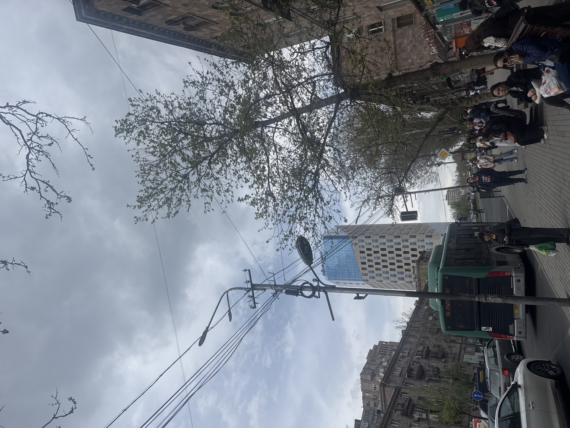 This image depicts a bustling urban street scene in Yerevan, Armenia, on an overcast day. The perspective is taken from a slightly upward angle, capturing a significant portion of the cloudy sky.
On the right side of the frame, a sidewalk is populated with several individuals. A group of people is gathered, with some seated on a bench under a tree, while others stand or walk. One person in a dark jacket is standing near the back of a green public bus, which is stopped on the road parallel to the sidewalk. The bus displays "HYUNDAI" and the license plate "N 5894" on its rear. Behind the bus, several cars, including a white and a silver vehicle, are visible on the road, alongside a blue circular street sign indicating a mandatory left turn.
Dominating the central foreground is a robust street light pole, from which numerous overhead wires extend across the frame. Buildings line both sides of the street. To the left, an older, multi-story building constructed of stone with numerous windows can be seen. Further down the street and to the right, a distinctive tall, modern high-rise building with a reflective blue glass facade and a grid pattern of square windows stands prominently against the sky. Other older buildings are visible further in the background.
Trees with budding green leaves and some bare branches are interspersed along the street, suggesting springtime. Commercial signs with various texts are visible on storefronts in the background, though most are unreadable due to distance; "TELLCELL" can be faintly discerned on one. The scene conveys the atmosphere of a typical daytime street in a city.