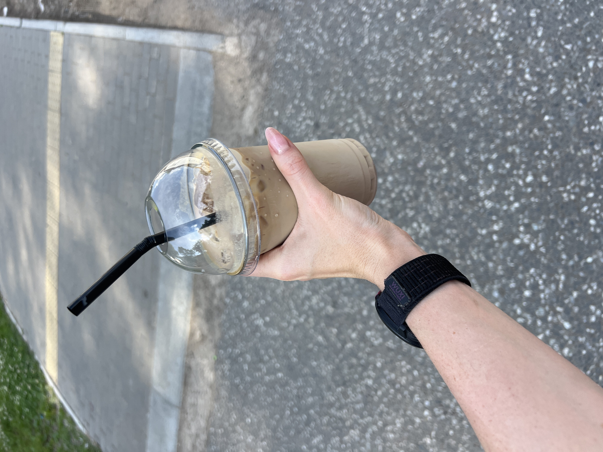 A person's left arm and hand hold a clear plastic cup filled with a light brown iced beverage and a black straw, angled slightly upward. The person is wearing a black watch on their wrist. The background is an outdoor paved surface, likely a sidewalk or pathway, with a strip of green grass and a concrete curb visible to the left. The setting is consistent with a public walkway in Chaikovsky, Russia, given the texture of the paving stones and the general urban park aesthetic.