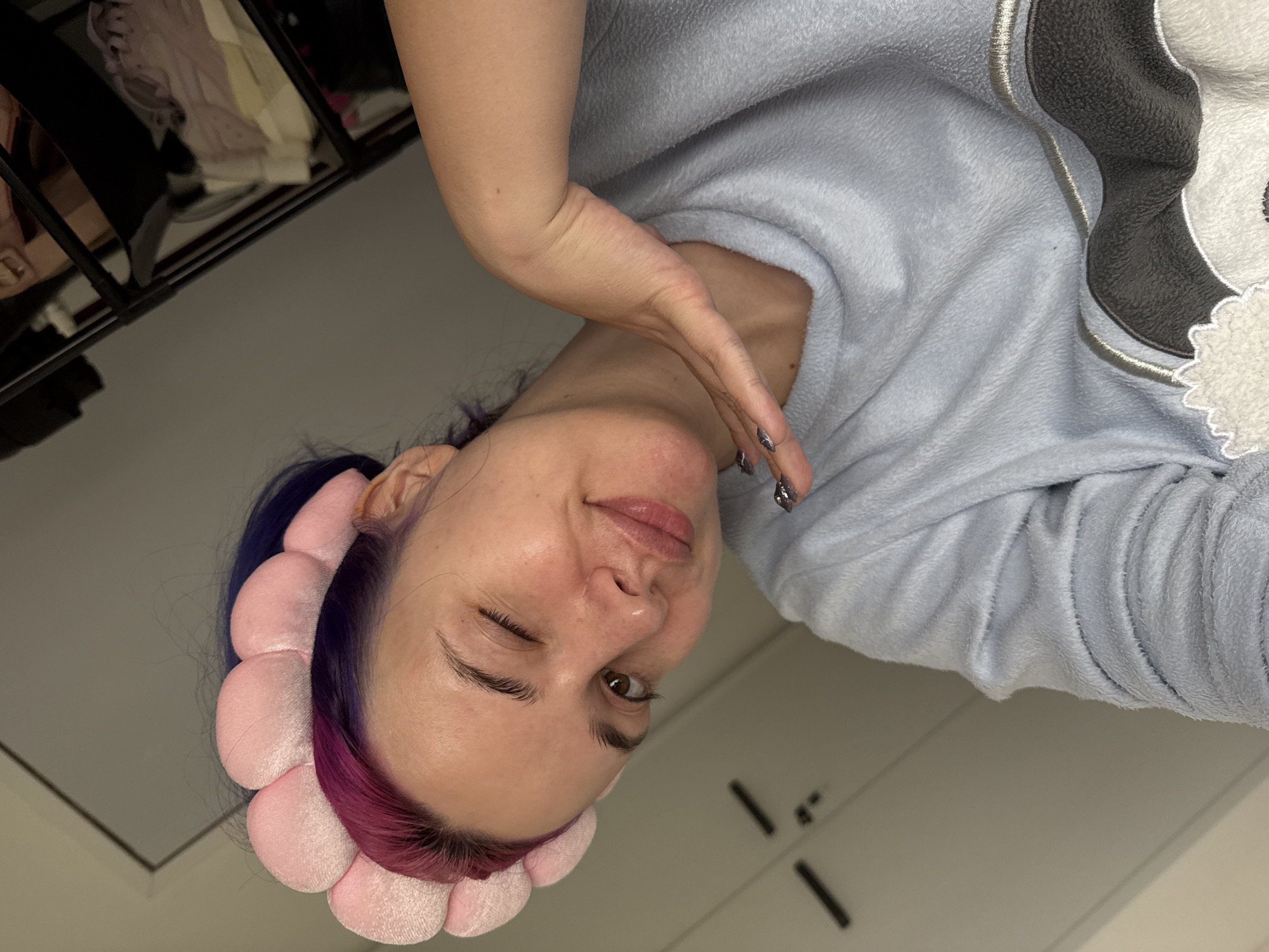 An indoor, rotated selfie-style portrait features a young adult with fair skin and striking dark purple hair that transitions to pinkish ends, held back by a plush, light pink, flower-petal-shaped headband. The person is wearing a cozy, light blue, fuzzy long-sleeved top adorned with a dark grey and white animal appliqué, possibly a panda or bear, on the upper right side. Their expression is playful, characterized by a wink from one eye and a slight smile, as they look directly at the camera. One hand is raised near their face, showcasing fingernails painted with a dark, possibly glittery silver, polish. Subtle skin imperfections are visible on the cheek and chin, giving a natural appearance.
The background includes a dark, multi-shelved unit on the left, likely a wardrobe or closet, with light-colored garments visible on its shelves. The rest of the background is a plain, light-colored wall or furniture surface, with dark, linear elements suggesting structural features or handles in the lower right. The overall setting is a personal indoor space, possibly a bedroom. The casual attire and headband suggest a relaxed moment, potentially during a self-care routine or leisure time. No text is visible in the image. The location context for this scene is the City of Zagreb, Croatia.