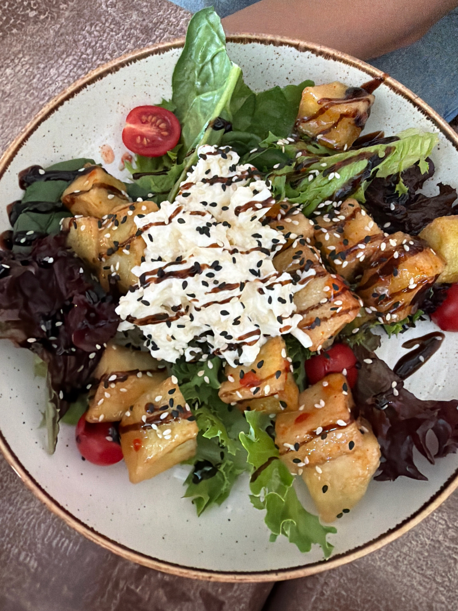 A close-up, overhead shot features a salad presented in a speckled, off-white ceramic bowl with a brown rim. The salad is composed of bite-sized, golden-brown fried pieces, possibly eggplant or tofu, glazed with a dark sauce and sprinkled with white and black sesame seeds. These pieces are arranged on a bed of mixed greens, including vibrant green lettuce and darker, reddish-brown leafy vegetables. Several halved cherry tomatoes are interspersed throughout the salad. At the center of the dish is a dollop of white, fluffy cheese or cream, also drizzled with the dark glaze and sesame seeds. The salad appears to be served on a textured, brown surface, likely a table. The background shows a blurred view of a person's arm and shoulder, indicating they are likely the one eating or preparing the meal. There is no visual information in the image to determine its location within Перовское сельское поселение, Ukraine.