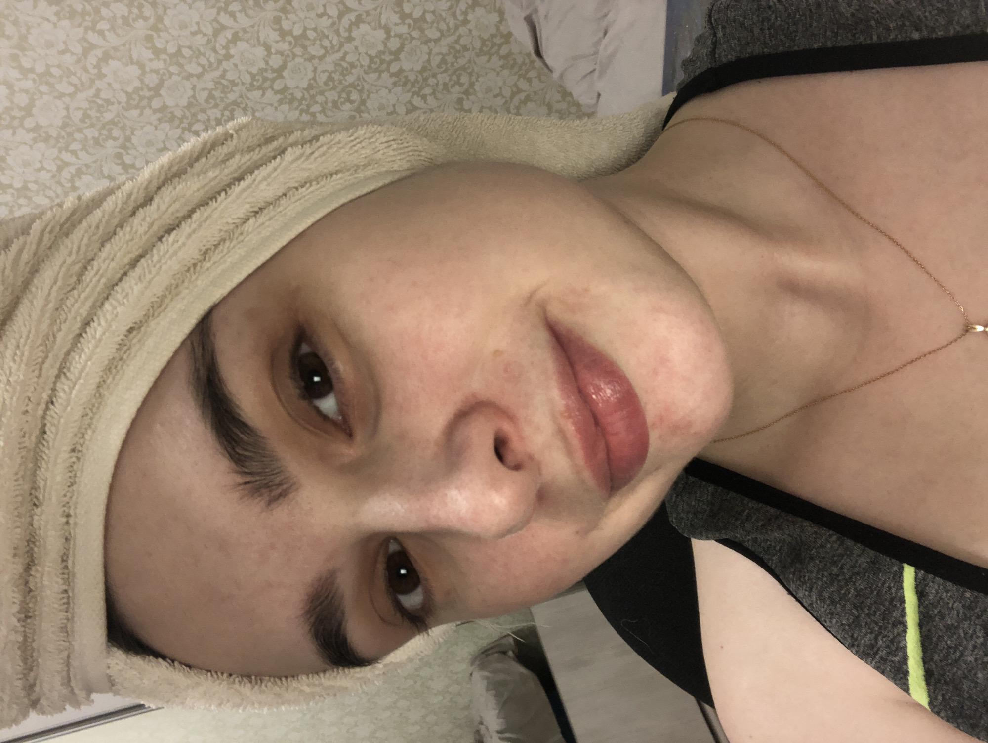 A person is seen in a close-up, head-and-shoulders shot, positioned at an oblique angle. The individual has fair skin, brown eyes, and dark eyebrows. A light-colored towel is wrapped around their head. They appear to be wearing a gray and black tank top with a thin, light green stripe. A delicate gold necklace is visible around their neck. The background consists of a patterned wallpaper with floral or damask motifs in muted tones. The location is unknown, and there is no indication of Noviy, Russia.