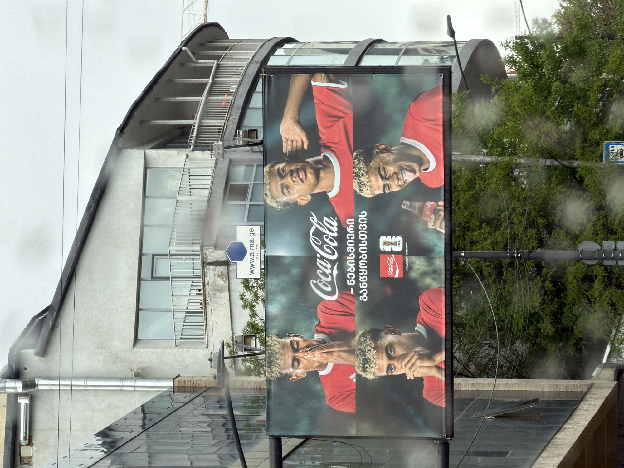 A billboard advertising Coca-Cola stands prominently in front of a building in Tbilisi, Georgia. The billboard features a repeating image of a young man in a red shirt, shown in a sequence of expressive poses. The Coca-Cola logo is prominently displayed, along with text in the Georgian language.
The billboard is situated in front of a building with a curved roofline and a series of windows. Additional architectural details include a metal staircase and various pipes or conduits running along the building's exterior. Lush green foliage, likely trees or shrubs, is visible behind the billboard. The overall environment suggests a possibly overcast day, with rain streaks on the camera lens.