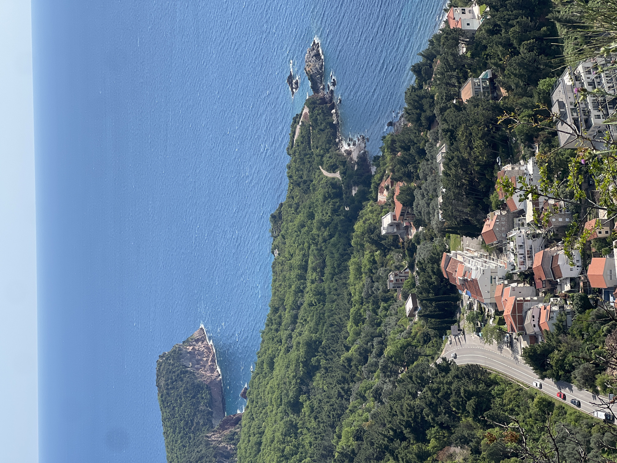 This high-angle, panoramic photograph captures a vibrant coastal landscape under bright, clear daylight. The scene, looking down over Sveti Stefan, Montenegro, features a picturesque blend of azure sea, verdant hillsides, and coastal architecture.
In the upper portion of the image, the vast, deep blue waters of the Adriatic Sea stretch towards a distant, hazy horizon, meeting a clear, light blue sky on the left. Several rocky formations and islets emerge from the sea, including a larger, vegetated island on the far left and a distinct, rugged rocky outcrop with a narrow, winding path extending into the water closer to the right.
The midground is dominated by a densely forested peninsula or headland, covered in lush green trees that cascade down towards the shoreline. Scattered within this greenery are numerous buildings, characterized by white walls and terracotta-tiled roofs, typical of Mediterranean architecture.
In the lower part of the frame, a winding, paved road snakes along the coastline, with several cars (including white, black, and red vehicles) visible on its surface. More buildings are clustered along this road and up the hillside, nestled amongst the abundant vegetation. The photograph is taken from a high vantage point, possibly a cliff or hill, with some tree branches partially framing the foreground on the right. The weather is sunny and serene, suggesting an ideal day for enjoying the scenic coastal environment. There is no visible text or clearly discernible people in the image.