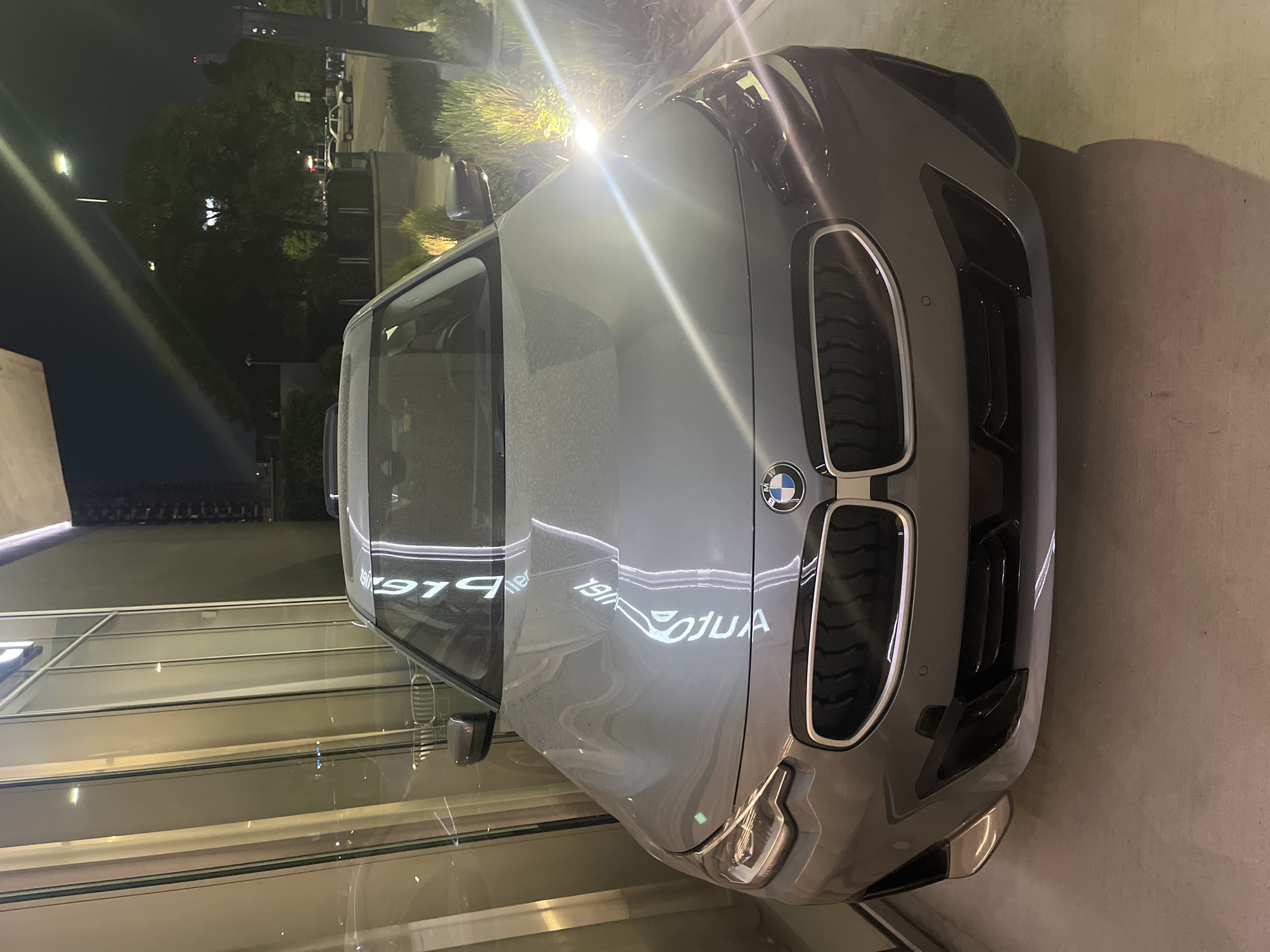 The image displays a gray BMW car parked at night, likely outside a dealership or showroom in Buenos Aires, Argentina. The car is viewed from a front-quarter angle, with its distinctive kidney grille and the BMW logo prominently featured.
The scene is illuminated by bright artificial lights, which create strong reflections on the car's polished hood and windshield. In the dark background, trees, streetlights, and other vehicles are dimly visible, indicating an urban or suburban setting. A powerful light source shines directly above the car's front.
Notable details include the mirrored text reflected on the car's surface: "AUTOLLER" and the number "1917" are clearly visible in reverse. These reflections likely originate from a sign on a nearby building, suggesting the car is on display at an automobile business. The reflections also show horizontal lines and interior lights from a large glass-fronted building. The car appears pristine, reinforcing the impression of a sales or display environment. No people are visible, and the primary activity is the static display of the vehicle. The time of day is clearly night, as evidenced by the dark sky and reliance on artificial lighting.