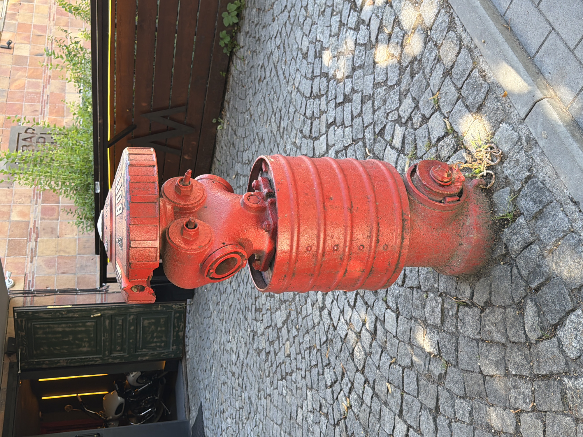 A red fire hydrant stands on a cobblestone sidewalk in front of an open doorway with a motorcycle visible inside. The hydrant is cylindrical with multiple nozzles and a cap on top. Behind the hydrant, a wooden fence with a metal "M" logo is visible. The background shows a brick building and green foliage. The scene is outdoors during daylight.