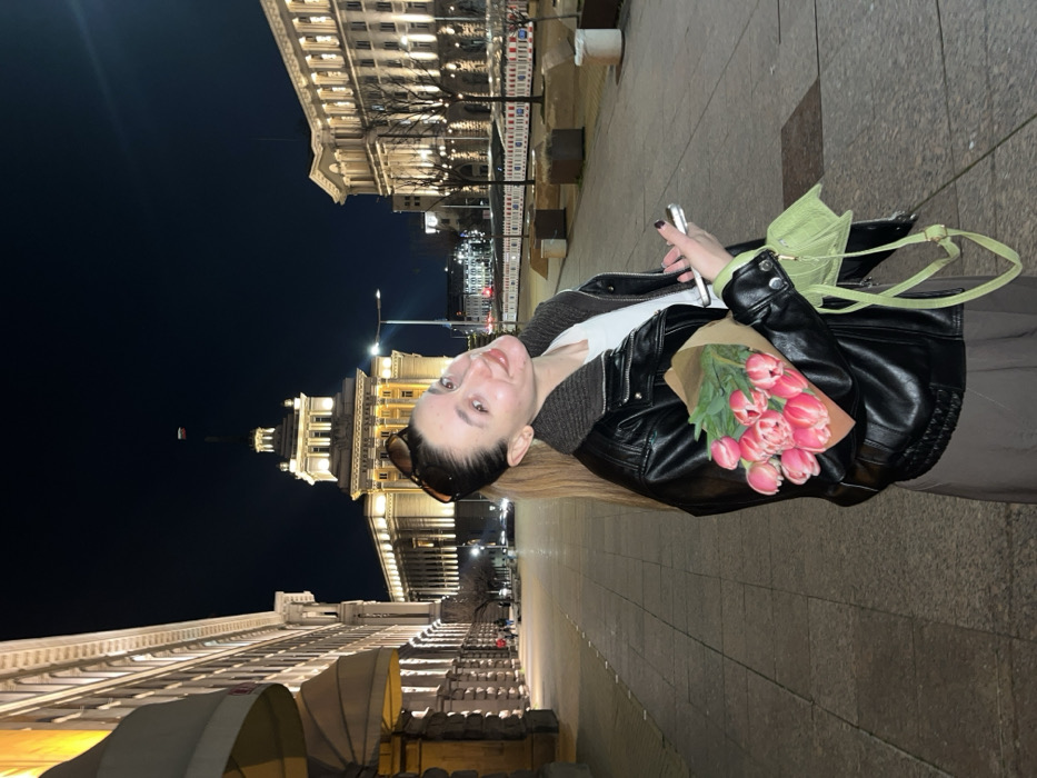 A young female stands smiling in an outdoor, nighttime urban setting, identified as the Largo complex in Sofia, Bulgaria. She has dark hair pulled into a high ponytail, with sunglasses resting on her head. She wears a black leather-style jacket with a ribbed grey collar, a white t-shirt, and dark trousers. In her left arm, she holds a bouquet of pink tulips wrapped in brown paper, while her right hand holds a silver smartphone. A small, light green cross-body bag hangs from her right side.
The background features the prominently illuminated Council of Ministers building (formerly the Party House), characterized by its central tower topped with a visible Bulgarian flag. To its right, the equally illuminated multi-story TSUM building (now office space) is visible. The scene is illuminated by artificial lights from the buildings and streetlights. The ground is a paved pedestrian area. Distant, blurred figures and vehicle lights are discernible further down the street.