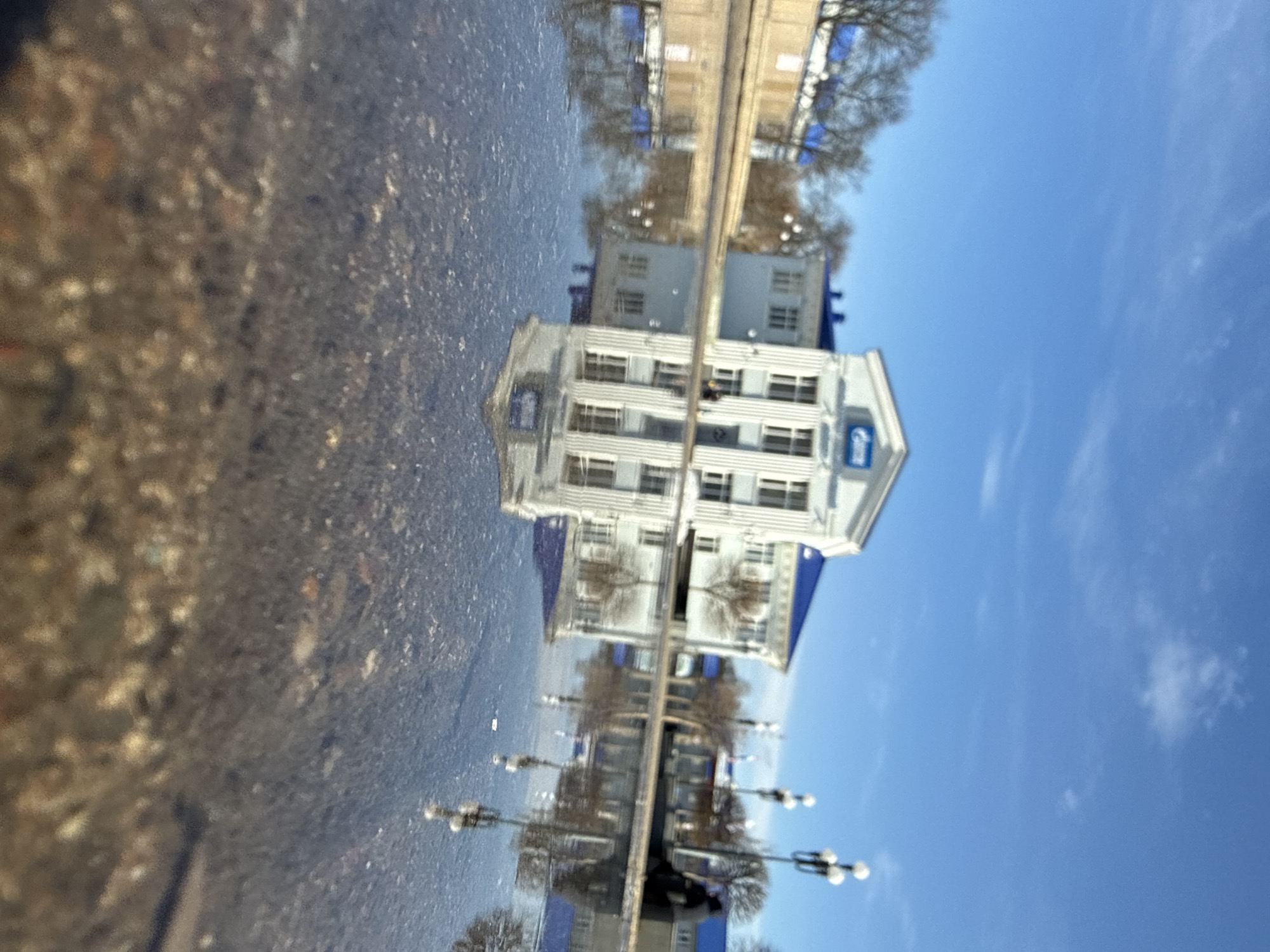 The image shows a reflection in a puddle. The reflection depicts a multi-story building with multiple windows and a sign that appears to read "Gazprom" (hard to confirm exactly due to reflection). The reflection also captures a street with trees and lampposts, and a blue sky with sparse clouds. The puddle itself shows the texture of the ground (possibly asphalt or concrete). The location is likely in a city that has a Gazprom building but the location name is not known.