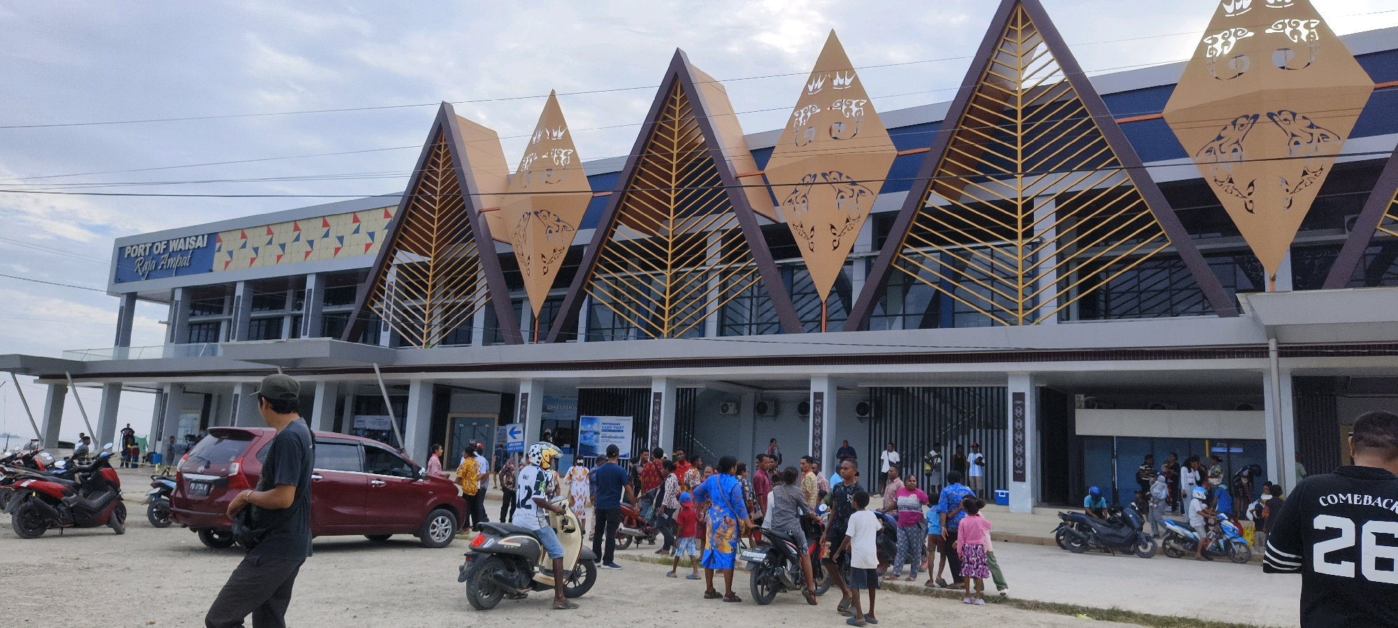 The image depicts the "PORT OF WAISAI Raja Ampat" building in Waisai, Indonesia, on a cloudy day. The modern building features a distinctive triangular roof design with gold-colored accents and intricate cut-out patterns. In front of the building, the ground is covered in a light-colored gravelly surface. A diverse group of people are present, some standing in clusters, others walking, and a few individuals are seated on motorcycles.
Several vehicles are visible: a red SUV is parked on the left side of the frame, and a maroon sedan is situated behind it. Various motorcycles are parked and being ridden by people. The people in the scene are dressed in casual attire, with a mix of bright colors and more subdued tones. Some individuals appear to be interacting with each other, while others are focused on their vehicles or moving through the area. The overall atmosphere seems to be one of a bustling public space, likely a transportation hub.