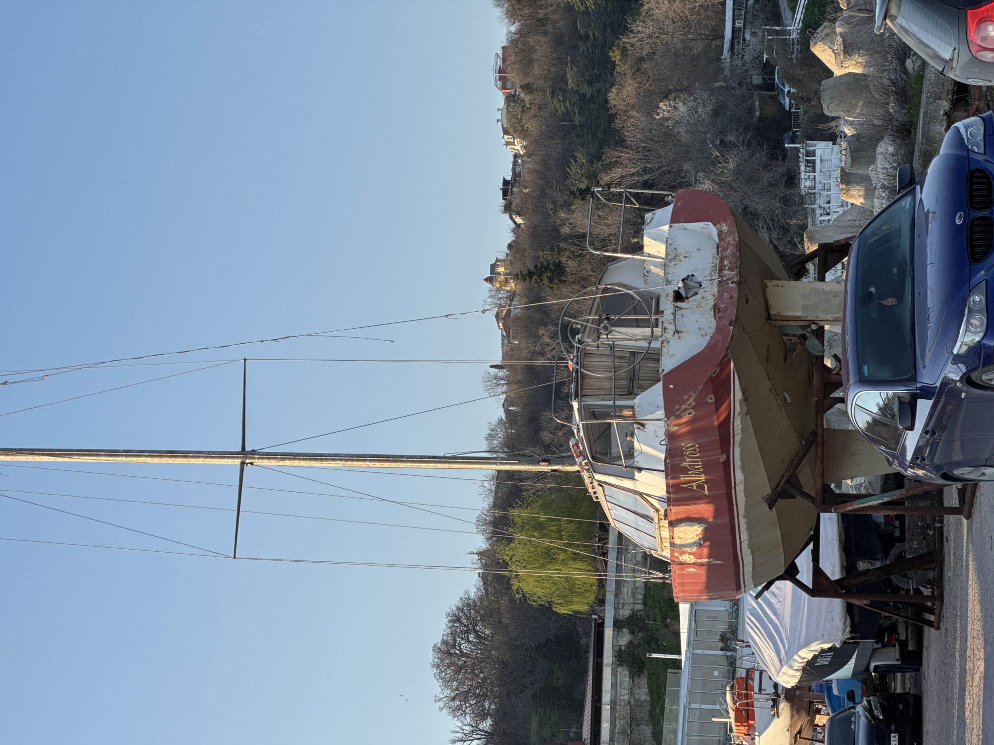 The image shows a docked sailboat named "Albatross" in Varna, Bulgaria. The boat, with a red and white hull, is supported on a metal frame on land, near a parked blue car. A tall mast with rigging extends upward. The background features buildings and trees on a hill. The sky is clear and blue.