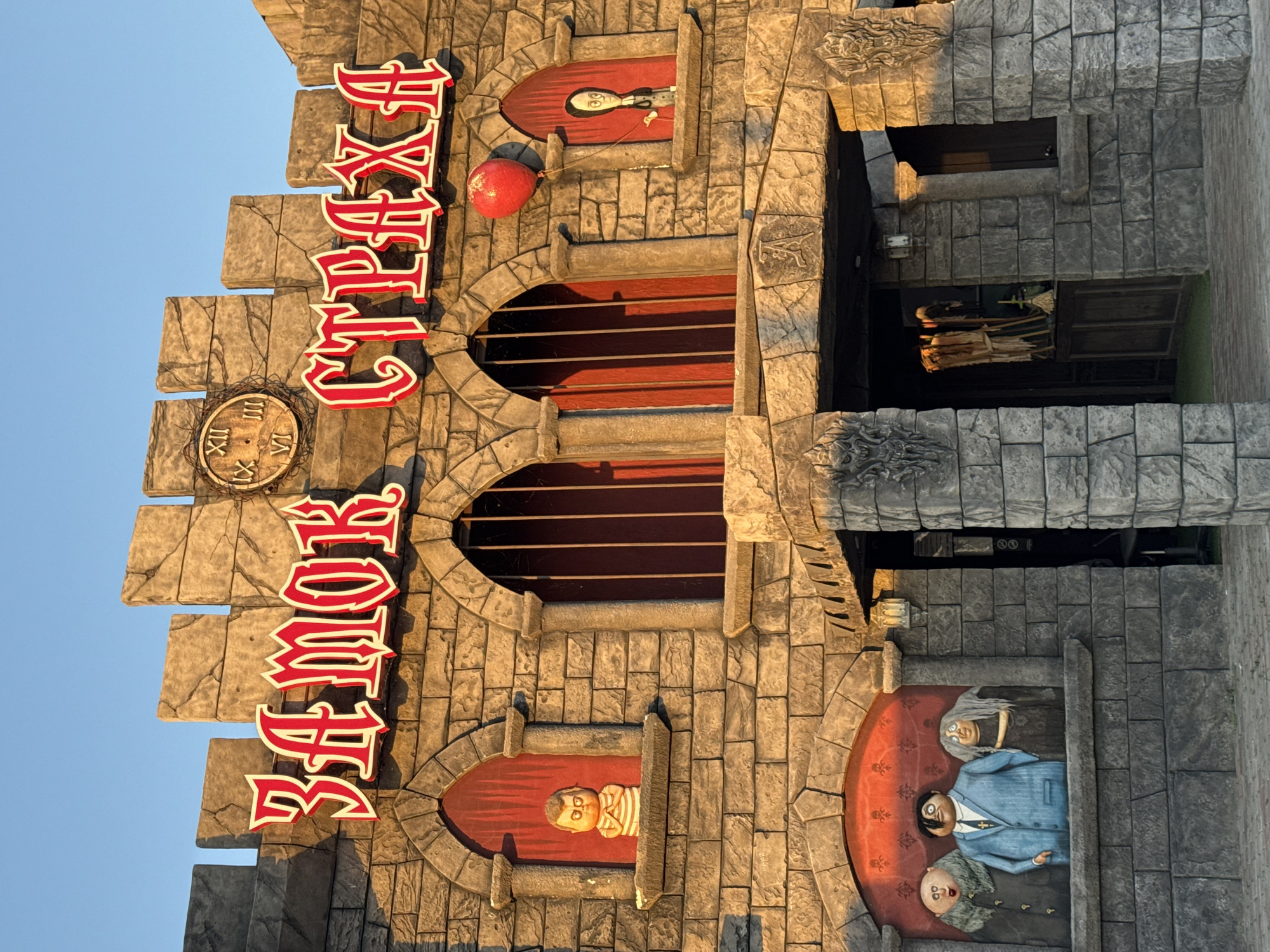 A gothic-style building facade, resembling a castle, is shown under a clear blue sky at sunset. The building is constructed with rough, grey stone blocks and features crenellated battlements along the top. Large, red, gothic-style lettering across the facade reads "ЗАМОК СТРАХА", which translates to "Castle of Fear".
To the left of the sign, within an arched window, a small, round-headed figure with striped clothing and bulging eyes is displayed. To the right of the sign, another arched window shows a slender, dark-haired figure in a black dress, holding a red balloon on a string. Above this figure, a large red balloon floats. A clock with Roman numerals is positioned above and between the two main windows.
Below the main sign, there are three figures depicted in a recessed alcove: a bald man with a fur collar, a man in a blue suit and tie, and an elderly woman with long grey hair. These figures appear to be characters from a fictional family, possibly inspired by the Addams Family.
The entrance to the building is located centrally, framed by stone columns adorned with carved grotesque faces. Inside the entrance, some decorative elements, including what appear to be straw figures and brooms, are visible. The building is located in Novofedorovka, Ukraine.
