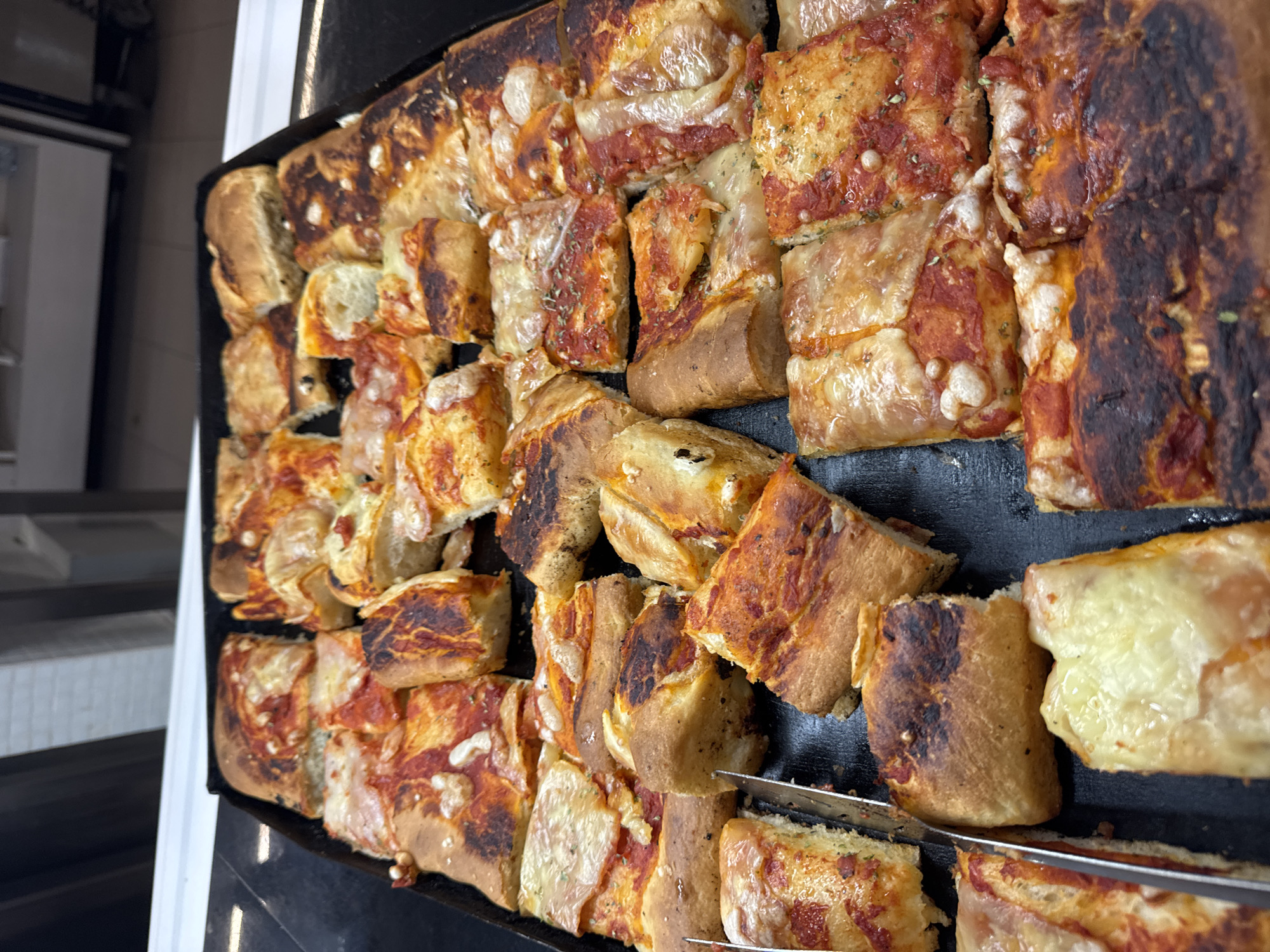 The image shows a close-up, high-angle view of a large tray filled with an assortment of rectangular pizza slices. The pizzas appear to be of a focaccia style, with thick crusts and various toppings. Some slices have a tomato sauce base with melted cheese, while others show more browning on the crust. In the foreground, on the left side, a pair of silver tongs are positioned over a slice of pizza, indicating that it is a self-service display, likely in a bakery or pizzeria. The background is out of focus but suggests an indoor setting with white walls and shelving. The overall impression is a visually appealing and abundant display of pizza. This scene is consistent with a food establishment in Isola delle Femmine, Italy, known for its culinary offerings.